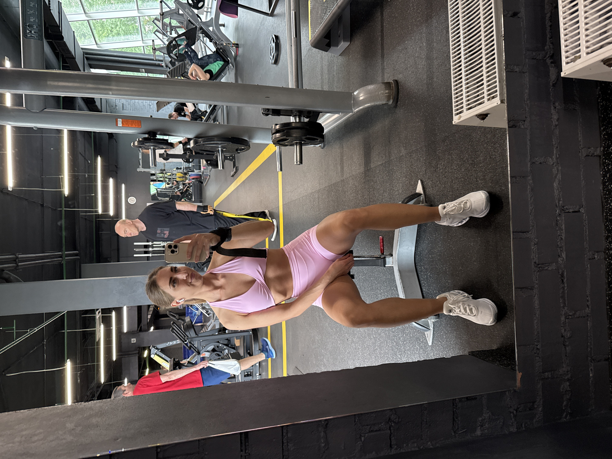A woman is taking a selfie in a gym. She is wearing a pink sports bra and pink shorts, and white sneakers. She is sitting on a weight machine. In the background, there are other people working out and gym equipment. The gym appears to be modern and well-equipped. The scene is illuminated by bright overhead lighting.