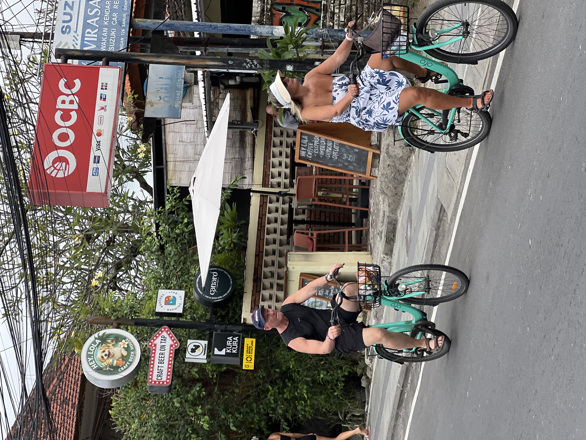 Two individuals are cycling on a street in Denpasar, Indonesia, on a cloudy day. The man, wearing a black sleeveless shirt, black shorts, and a blue baseball cap, rides a turquoise bicycle with a basket containing items. The woman, in a strapless blue and white floral dress and a beige sun visor, rides a similar turquoise bicycle, also with a basket. They are positioned on the road, with a sidewalk and buildings visible behind them. Signs for "HEY LOLA," "CRAFT BEER ON TAP," and "OCBC" are prominent in the background, suggesting a commercial area. The environment appears urban, with overhead wires, trees, and buildings indicating a street scene.