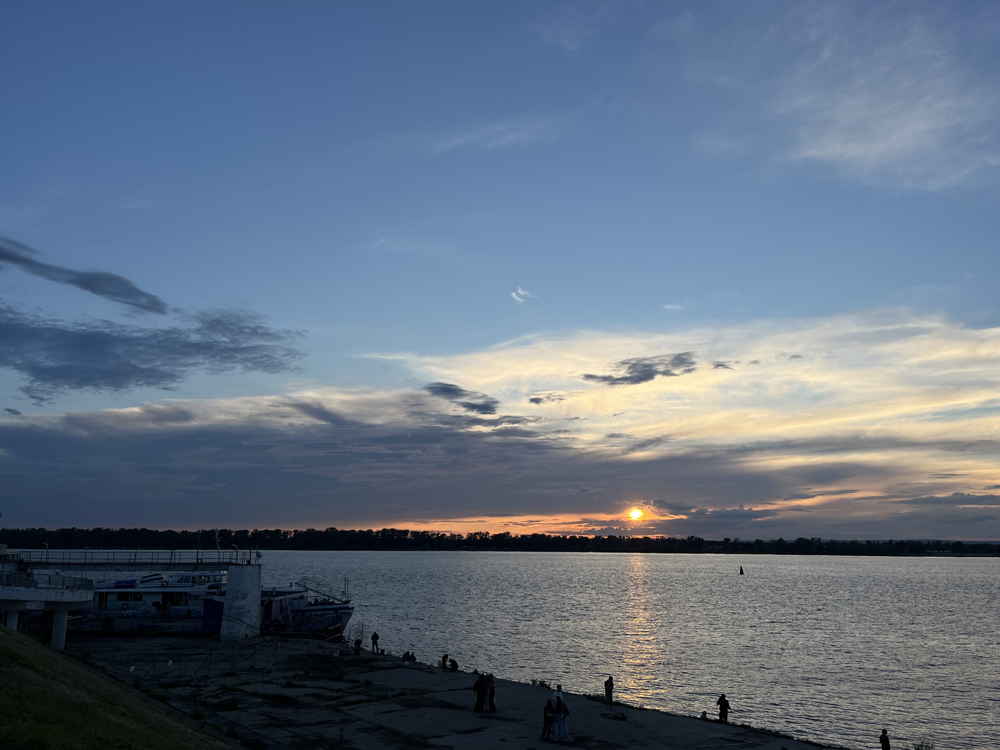 The image depicts a scenic sunset over a wide river, likely the Volga River, near Novokuybyshevsk, Russia. The sky is a gradient of blues and yellows, with dramatic clouds reflecting the warm hues of the setting sun. The sun itself is a bright orb on the horizon, casting a shimmering reflection on the water.
On the riverbank in the foreground, a concrete embankment is visible, with several people scattered along it, some appearing to be fishing or simply enjoying the view. A few boats are docked along the embankment, suggesting a populated riverfront. The far bank of the river is lined with a dense forest, silhouetted against the colorful sky.
The overall mood is peaceful and serene, evoking the quiet beauty of a summer evening. The time of day is clearly sunset, given the low angle of the sun and the warm, golden light. The weather appears calm, with light clouds scattered across the sky. There is no visible text in the image.