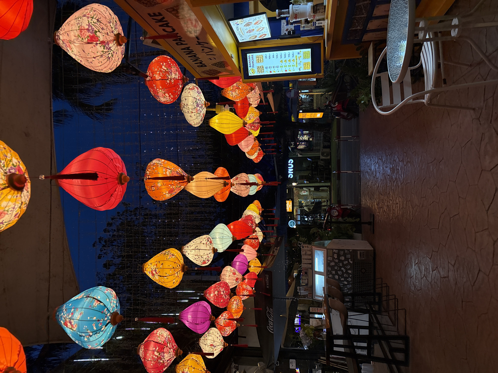 A street scene in Choeng Thale, Thailand at night, featuring a row of colorful illuminated paper lanterns hanging overhead. The lanterns, in various shapes and patterns, are predominantly red, pink, yellow, blue, and white, many adorned with floral motifs. Below the lanterns, a pathway is paved with textured red-brown tiles. On the right side of the image, a yellow food stall with signage for "BANANA PANCAKE" is visible, displaying menus with prices and food items. A white metal table and chair are situated in front of the stall on the pathway. To the left, a bar area with stools and a Coca-Cola umbrella is partially visible. Further down the street, other shops and buildings are discernible, along with trees and streetlights. The overall ambiance is festive and commercial.