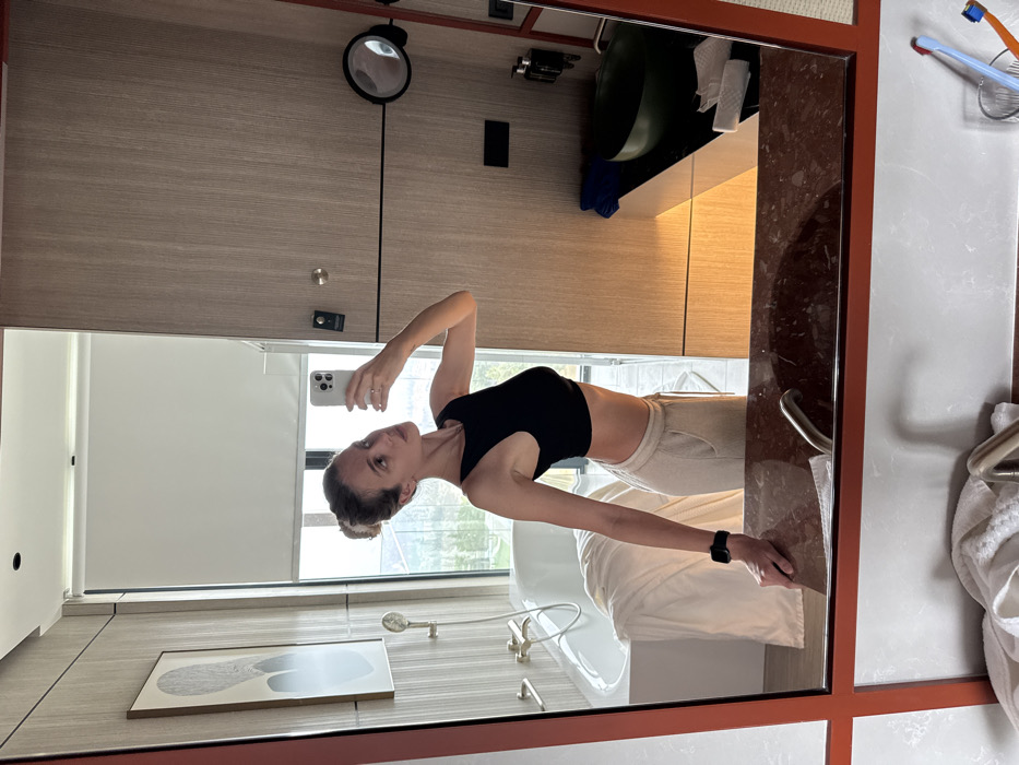 The image displays a selfie taken by a woman reflected in a large mirror within a modern, well-appointed hotel bathroom, strongly suggesting a location within the Rooms Hotel Tbilisi in Tbilisi, Georgia, based on the distinctive interior design elements and visible city view.
The central subject is a woman, appearing in her 20s or 30s, reflected from the waist up. She has light skin and her hair is pulled back into a bun. She wears a black sleeveless crop top and light-colored, loose-fitting pants. A dark smartwatch is visible on her left wrist. She is holding a light-colored smartphone in her right hand, positioning it to capture the reflection. Her left arm rests on the dark reddish-brown marble countertop of the vanity. She is looking slightly upward and to her left, towards the phone's camera.
The bathroom features light wooden paneling on the walls and a contemporary aesthetic. To the left of the woman's reflection, a white, modern bathtub with gold-toned fixtures and a handheld showerhead is visible. A white towel or robe is draped over the tub's edge. A large window with a partially raised white roller blind overlooks a landscape of green hills and distant structures, characteristic of Tbilisi's elevated views. A framed abstract artwork hangs on the wall above the tub. To the right of the woman, the reflection shows a dark green, bowl-shaped sink set into a dark countertop, equipped with a brass-colored faucet and wall-mounted dispensers. A circular magnifying mirror with a black frame is attached to the wall. Paper towels and a blue towel are present on this countertop. In the foreground, below the mirror's frame, the actual sink area shows a metal faucet, a white towel, and a clear cup holding a blue and an orange toothbrush.