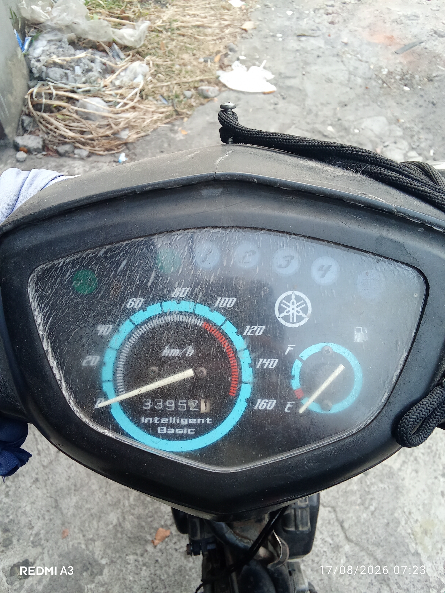The image shows a close-up view of a motorcycle's instrument cluster. The odometer reads 33952, and the text below it says "Intelligent Basic." The speedometer has markings up to 160 km/h, with the needle currently indicating approximately 20 km/h. A fuel gauge shows the fuel level to be between empty (E) and full (F). There are indicator lights for neutral, 1st gear, 2nd gear, 3rd gear, and 4th gear. A Yamaha logo is visible in the center of the cluster. The instrument cluster is dusty. The background shows a patch of dry grass and some debris, and a concrete surface. The timestamp in the lower right corner indicates the image was taken on August 17, 2026, at 07:23. The text "REDMI A3" in the lower left corner suggests the device used to capture the image. The location is identified as Sanggrahan, Indonesia.