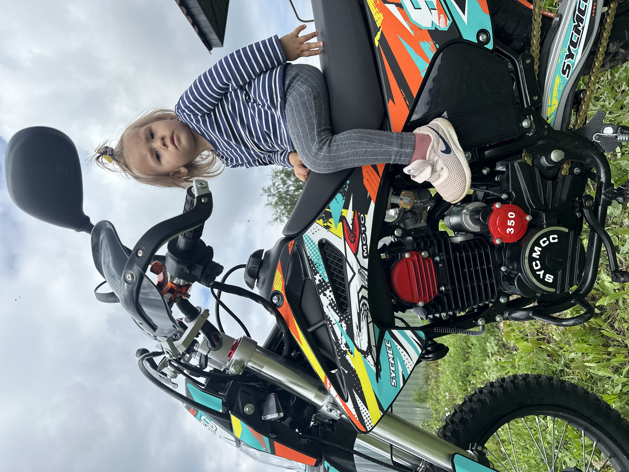 A young child sits on the seat of a dirt bike outdoors on a cloudy day. The child has blonde hair pulled into a ponytail and is wearing a blue and white striped long-sleeved shirt and gray striped pants. The child is wearing light pink sneakers. The dirt bike is black with colorful graphics in orange, teal, yellow, and white. The child's feet are resting on the engine block of the motorcycle. The background shows green grass and some trees, with a cloudy sky above. The scene appears to be in a rural or outdoor setting, possibly a yard or field. There are no visible buildings that would definitively place the location within Kharnavy, Russia.