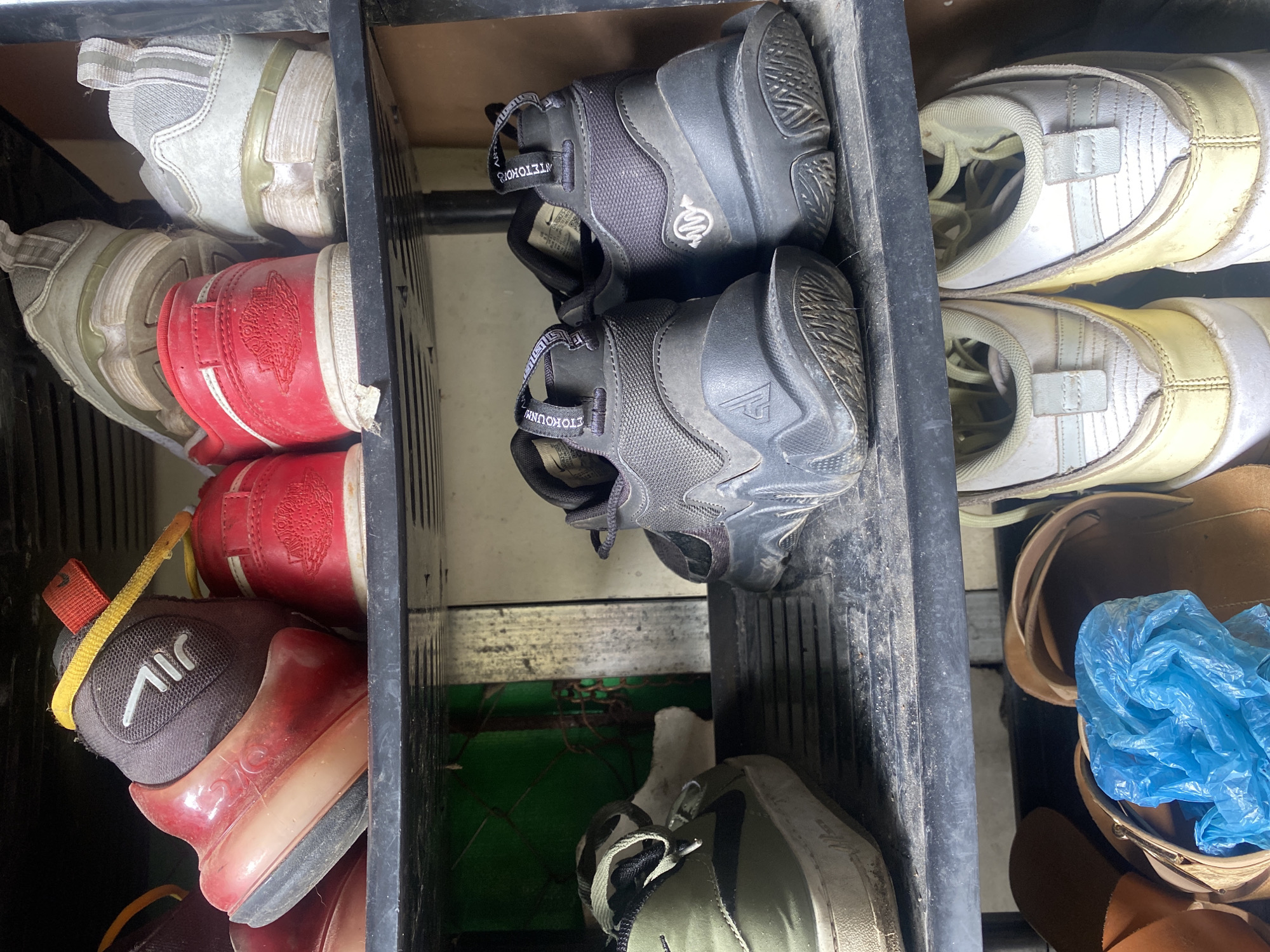This media file captures a functional shoe storage setup, likely situated in an entryway or outdoor area in Davao City, Philippines. A dark, multi-tiered shoe rack is visible, filled with various worn pairs of shoes, mostly oriented with their heels facing outwards.
On the upper left shelf, a light-colored athletic shoe rests next to a red and white shoe, which displays an embossed circular design on its heel, indicative of Nike Air Jordan branding. The middle shelf features two black Nike Zoom Freak sneakers, identifiable by the "ANTETOKOUNMPO" text on their pull tabs and a distinct logo on the heel. Adjacent to these, on the upper right side, are two light yellowish-white athletic shoes with horizontal stripes and a Velcro strap.
The lower left section of the rack contains a dark grey shoe with a red translucent sole, marked with "JIL" text and a white swoosh-like emblem. Another red shoe is partially visible beside it. A green, possibly olive-colored, shoe with white laces is also present. In the bottom right, a brown sandal or slipper is seen, containing a crumpled blue plastic bag.
The shoes appear well-used and dusty, suggesting regular wear. The bright lighting indicates it is daytime. A green mesh or fence-like structure is visible in the background, reinforcing the impression of a semi-outdoor or utilitarian setting common in local households for shoe storage.