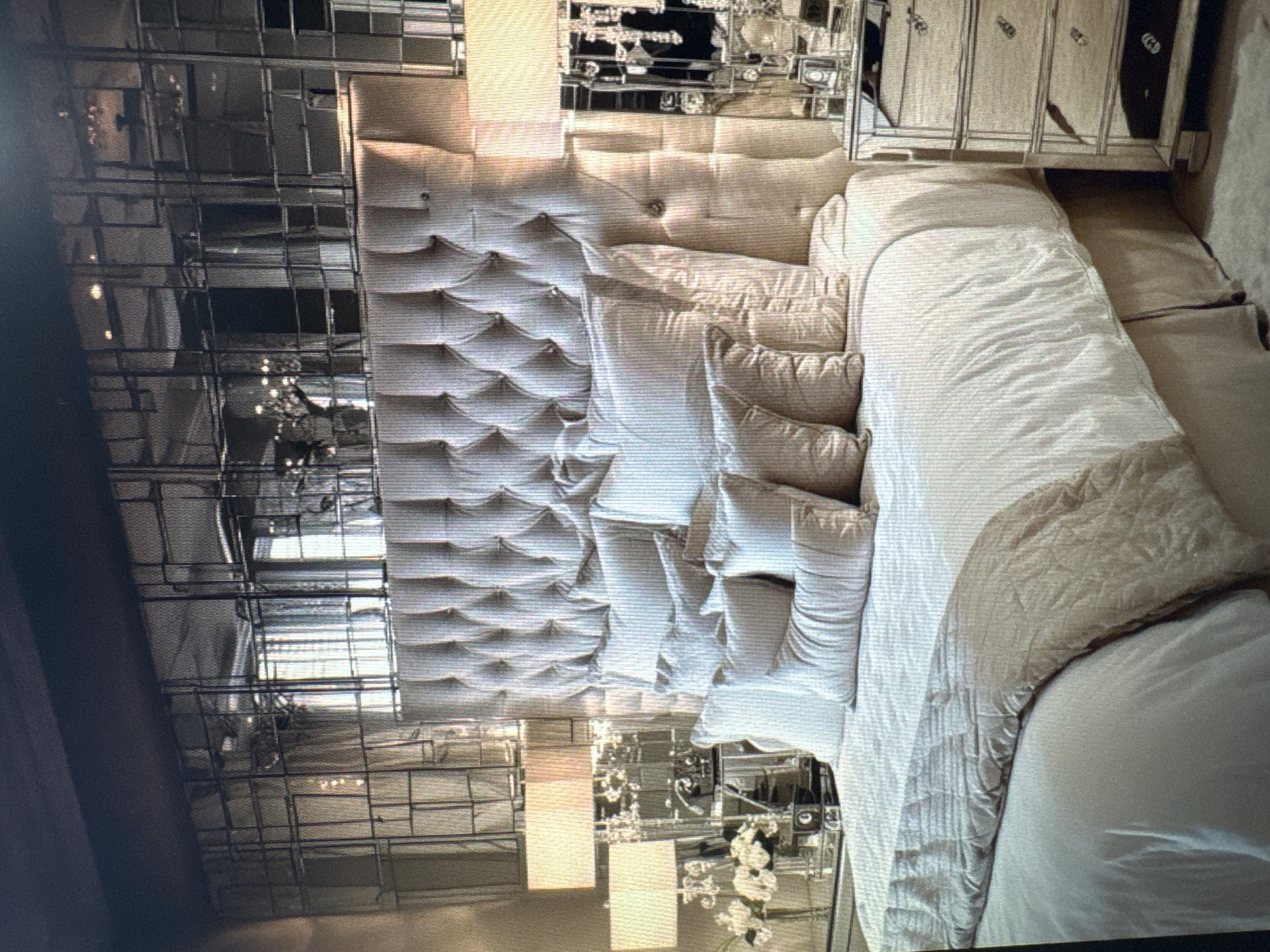 This media file shows a bedroom with a large, tufted headboard and a bed covered in white linens and pillows. To the left of the bed is a tall, mirrored furniture piece with multiple shelves and drawers. To the right of the bed, more mirrored furniture, possibly a dresser or wardrobe, is visible, featuring several drawers and decorative hardware. The headboard is a prominent feature, with a diamond-tufted pattern that creates a textured, upholstered look. Several pillows are arranged on the bed, some matching the headboard in color and texture, while others are a lighter shade of white. A decorative throw or duvet, in a neutral tone with a subtle pattern, is draped across the foot of the bed. The overall aesthetic suggests a luxurious and sophisticated bedroom. The presence of mirrored furniture and the extensive use of light colors contribute to a bright and airy feel, reflecting light throughout the space. The background appears to be a wall, also with some reflective elements. The image is an interior shot, likely taken during the day given the lighting. No people are visible, and no specific time of day or weather conditions can be inferred beyond indoor lighting. No text is visible in the image. The location context of Ellwangen, Germany does not offer any specific visual cues within the image itself.