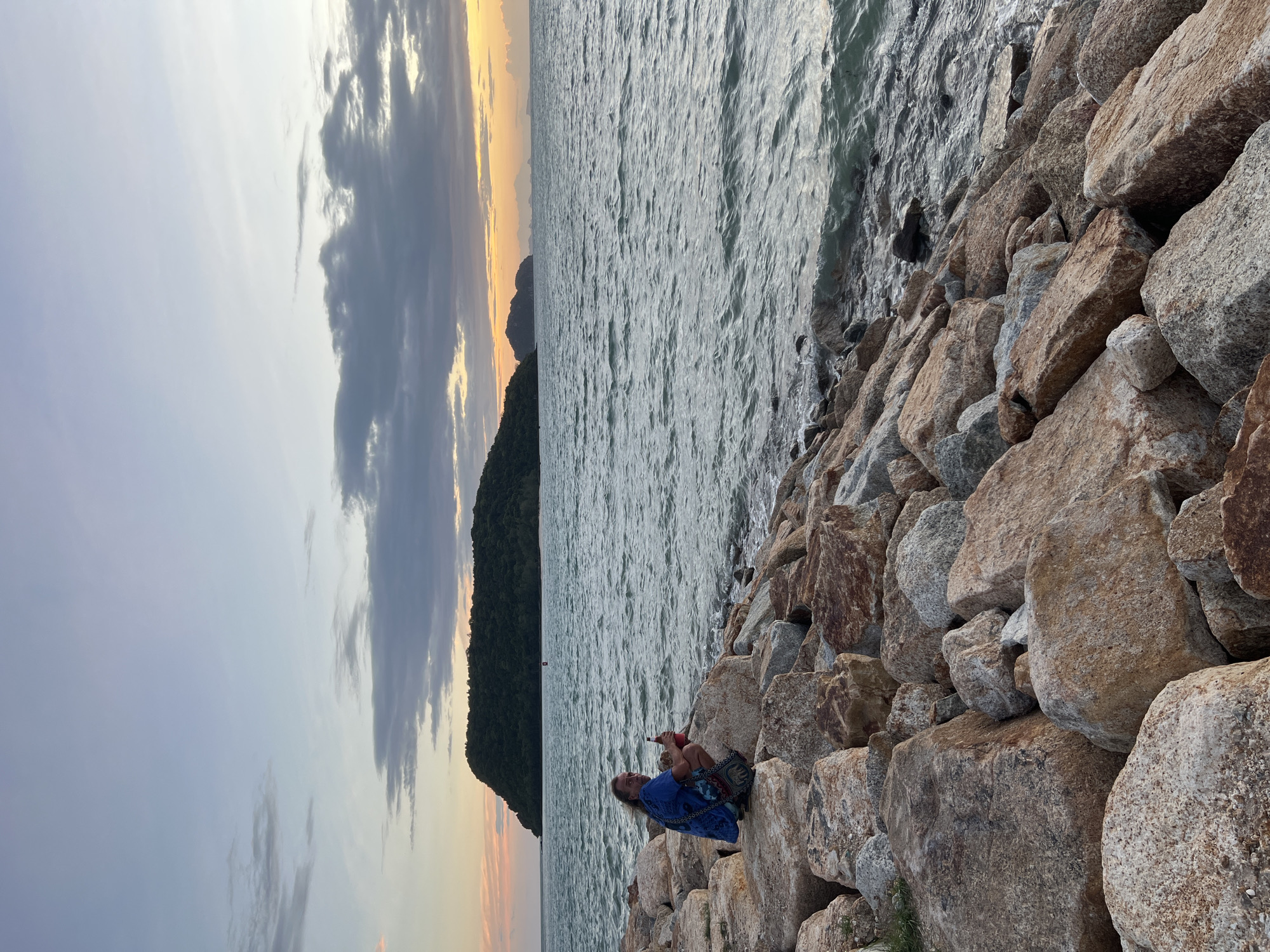 A man with long hair, wearing a blue shirt and a patterned bag, crouches on a rocky shore overlooking the ocean. He holds a bottle in his hand. The sky above is a gradient of pale blue and orange, with clouds scattered across it. In the distance, a lush green island is visible. The sea is choppy, with waves lapping against the rocks. The scene appears to be set in Ko Pha-ngan District, Thailand, during sunset or sunrise.