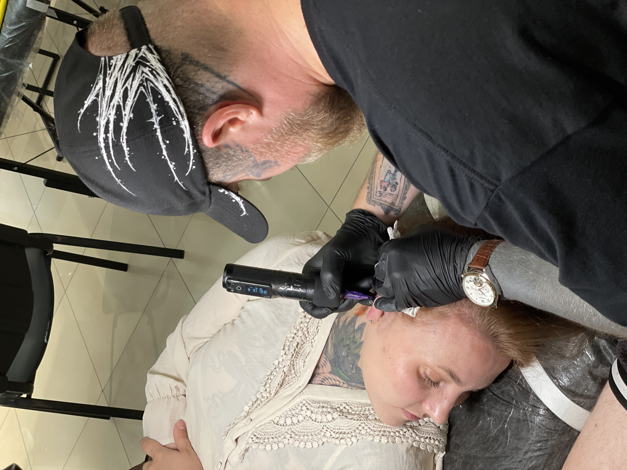 An overhead, medium shot captures a tattoo artist in black gloves meticulously working on a client's face in an indoor setting. The client, lying down with eyes closed and wearing a cream-colored blouse, is positioned on the left side of the frame. The artist, seen from the back and right shoulder, is wearing a black baseball cap with white abstract designs, a black t-shirt, and a brown leather-strapped watch on their left wrist. The artist holds a black tattoo pen with a purple tip in their gloved right hand, applying it to the client's eyebrow area. The floor, tiled in a beige and cream pattern, is visible in the background, along with the legs of a dark chair. A tattoo is partially visible on the client's chest and arm.