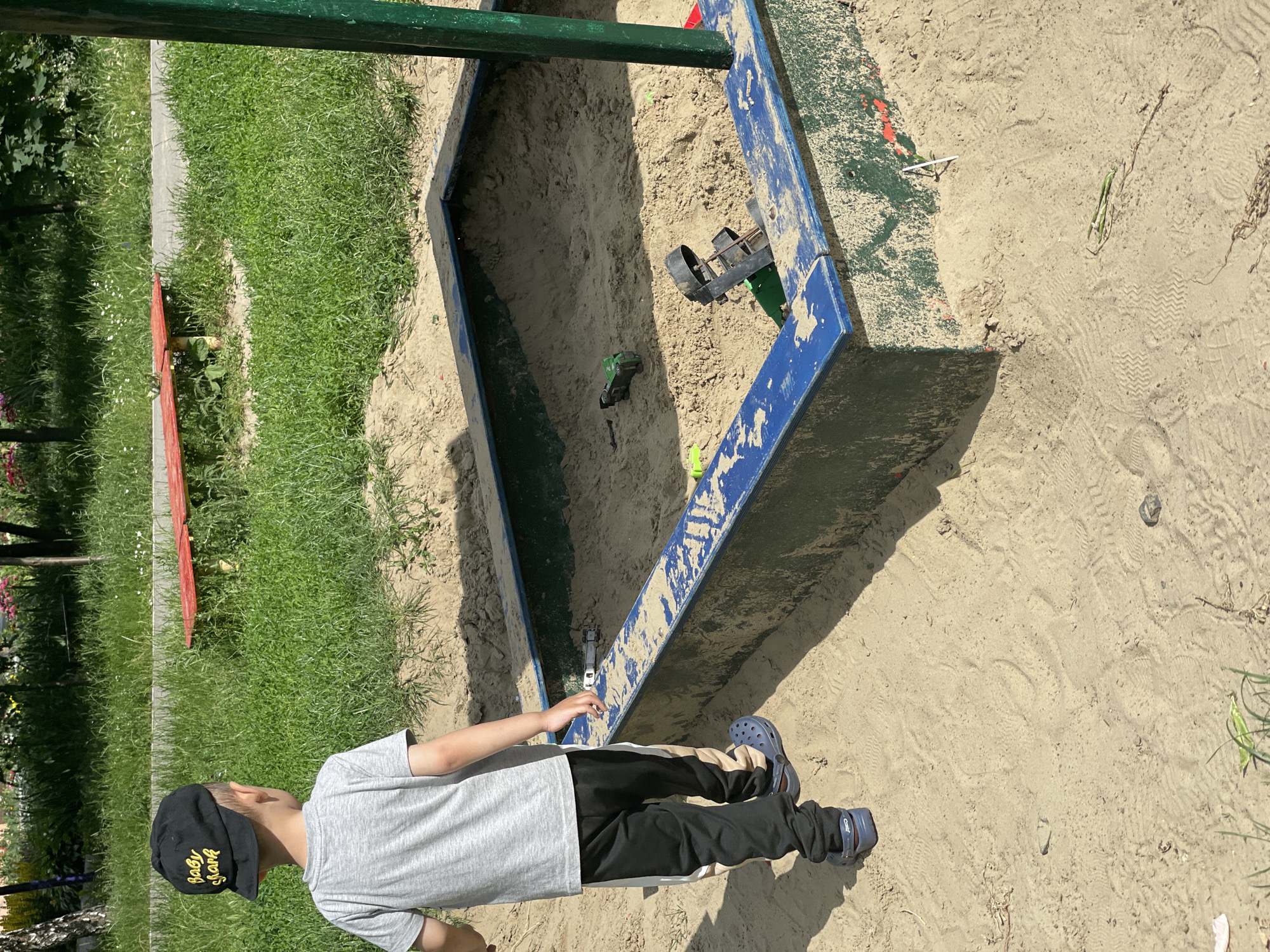 An outdoor, eye-level, full shot captures a young boy wearing a black baseball cap and a gray t-shirt standing in a sandbox. The sandbox, with blue trim and green sides, is filled with sand and toys, including two green toy trucks. To the left of the sandbox, the boy's legs are visible from the knees down, wearing black pants with a beige stripe and blue crocs. Behind the boy, a patch of green grass and a red bench are visible. The sand in the sandbox shows footprints.