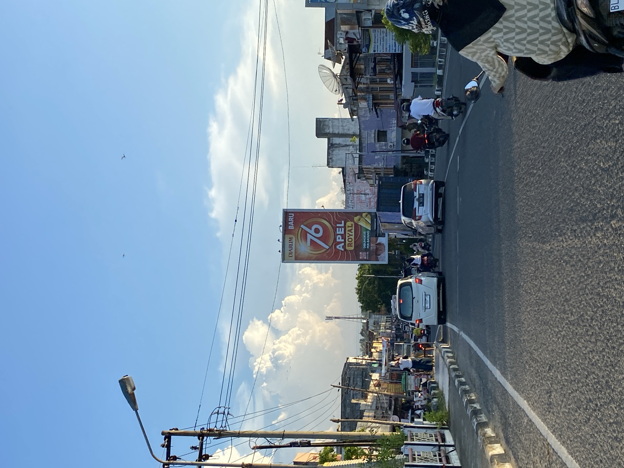 The image depicts a street scene in Lhokseumawe, Indonesia, under a clear blue sky with scattered white clouds. The perspective is from a moving vehicle, likely a motorcycle, looking forward. The street is paved and has a white line marking the lanes.
On the right side of the road, there is a woman riding a motorcycle in the foreground, wearing a patterned hijab and a helmet. Further down the road, two men on motorcycles are visible, one in a white shirt and the other in a maroon shirt, both wearing helmets.
On the left side of the road, there is a sidewalk with a white and black curb. Several people are standing or walking on the sidewalk, some near food stalls or shops. A utility pole with wires runs along the sidewalk.
In the background, there are buildings lining the street, varying in architectural style and condition. A large billboard advertising "76 APEL ROYAL" is prominently displayed. Other smaller signs and satellite dishes are visible on the rooftops of some buildings. Trees and greenery are also present in the distance, suggesting a mix of urban and natural elements. The lighting suggests it is late afternoon or early morning, with the sun casting long shadows. Two birds are visible in flight against the sky.