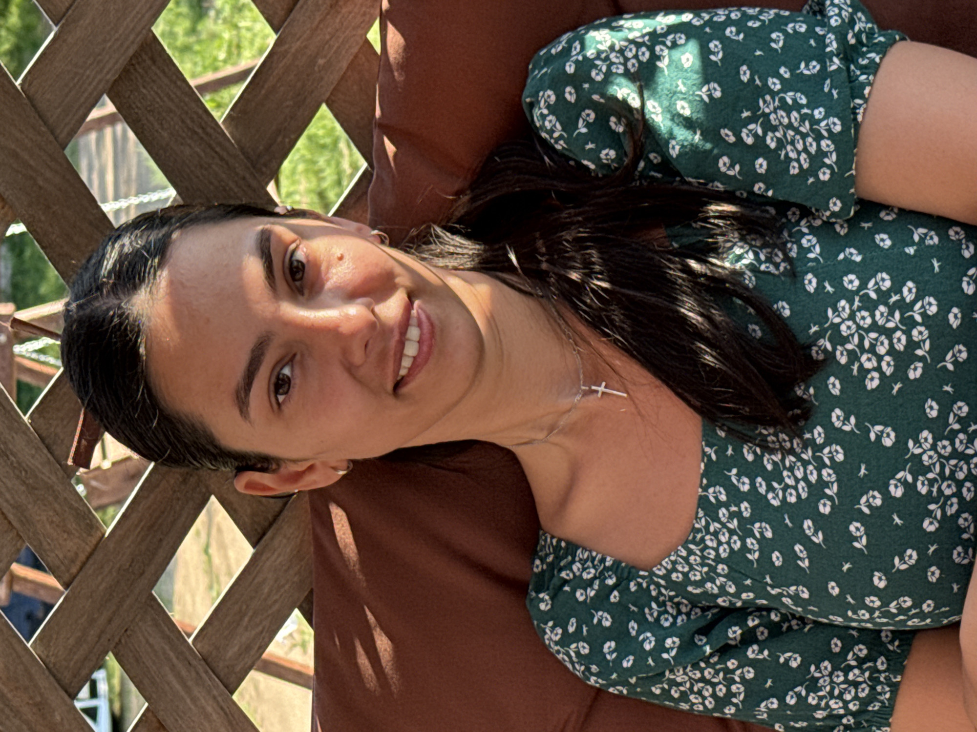A young woman lies on a brown, cushion-like surface, possibly a lounger, outdoors. She is smiling and facing the camera. She has dark hair, light skin, and is wearing a green dress with a white floral pattern and a silver necklace. Part of a wooden lattice or fence is visible to the left, with foliage seen through the openings. The setting appears to be sunny. It is unknown if this image was taken in Chaikovsky, Russia.