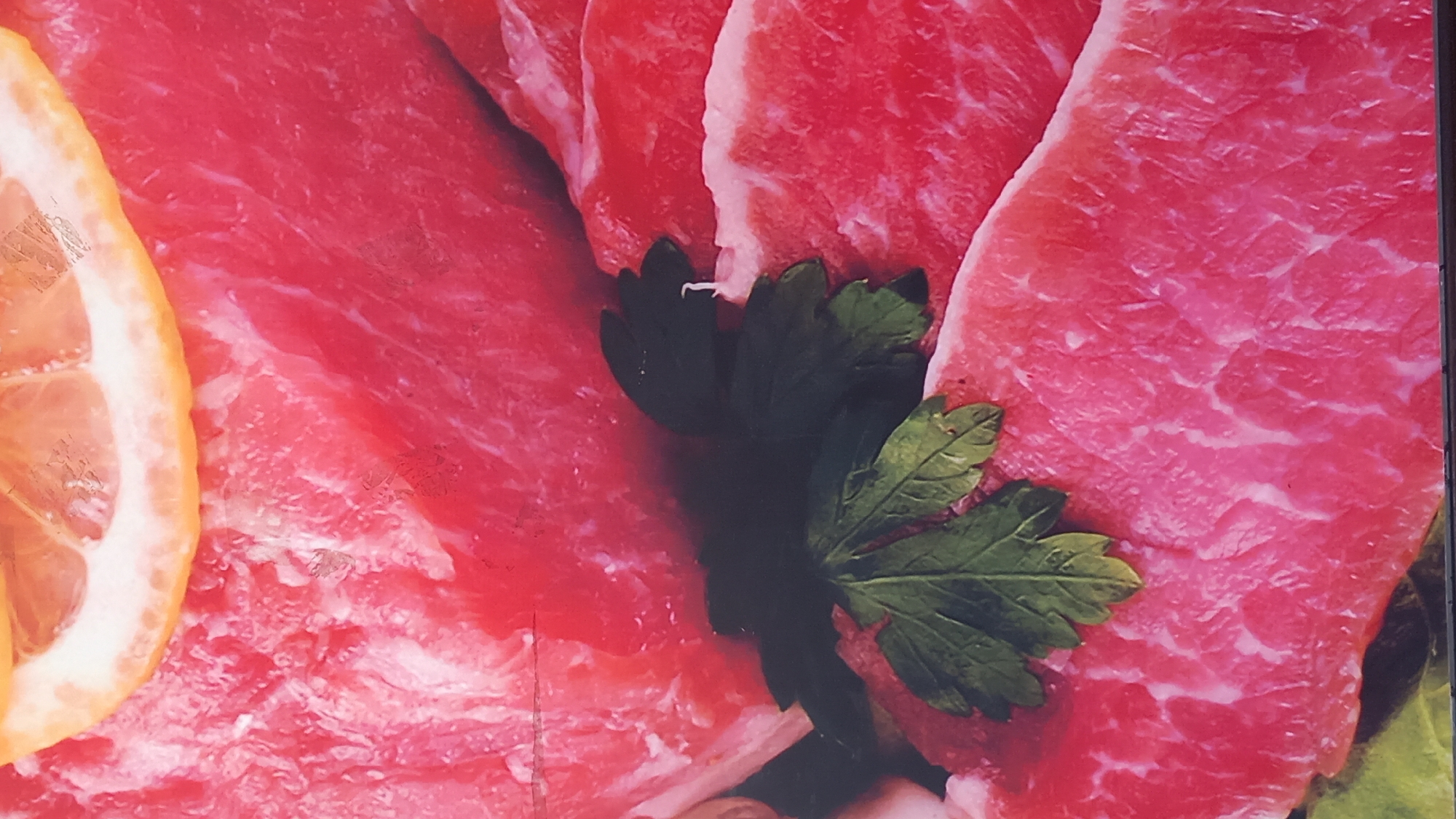 The image displays raw red meat, likely beef steaks, with visible marbling and a sprig of green parsley. A thin slice of lemon is positioned at the top of the frame, resting on the meat. The background is a blurred texture of more raw meat. There are no people or visible structures that would indicate a specific location in the city of Kos, Greece. The lighting suggests an overhead light source. The focus is on the food items themselves, with the parsley and lemon providing a decorative element to the presentation of the meat. The overall impression is that of a food photograph or advertisement.