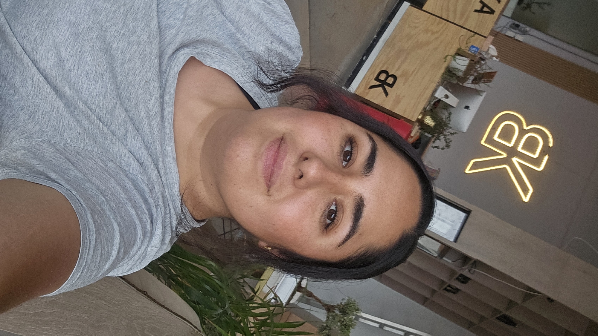 A woman poses for a selfie in an indoor setting, likely a café, indicated by the illuminated "KB" neon sign and wooden décor elements in the background. She is wearing a grey t-shirt. A plant is visible in the foreground. The location is potentially in Puebla, Mexico.