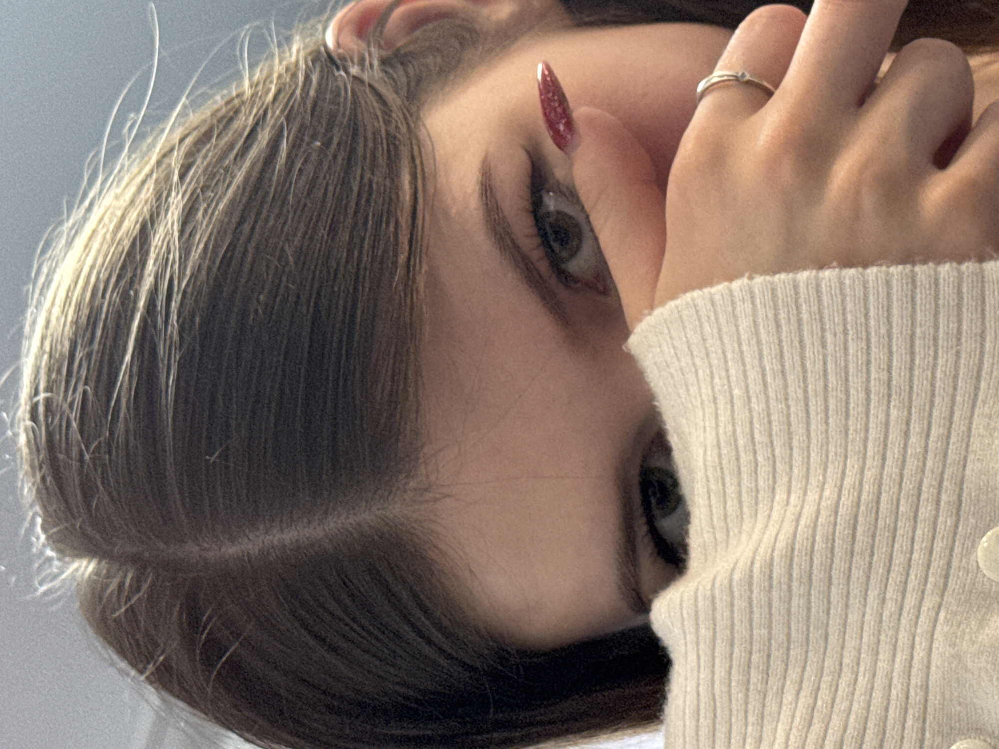 The image is a close-up, overhead shot of a young woman's face, partially obscured by her hand. She has brown hair, styled with a center part, and is wearing a cream-colored sweater. Her right eye is visible, with dark eyeliner and a winged flick. Her fingernail on the same hand is painted a sparkly red. A silver ring is on her ring finger. The background is out of focus and appears to be a light-colored, textured surface. The lighting suggests it is daytime, possibly indoors near a window. The overall tone of the image is casual and personal. The location is not specified in the image or its metadata.