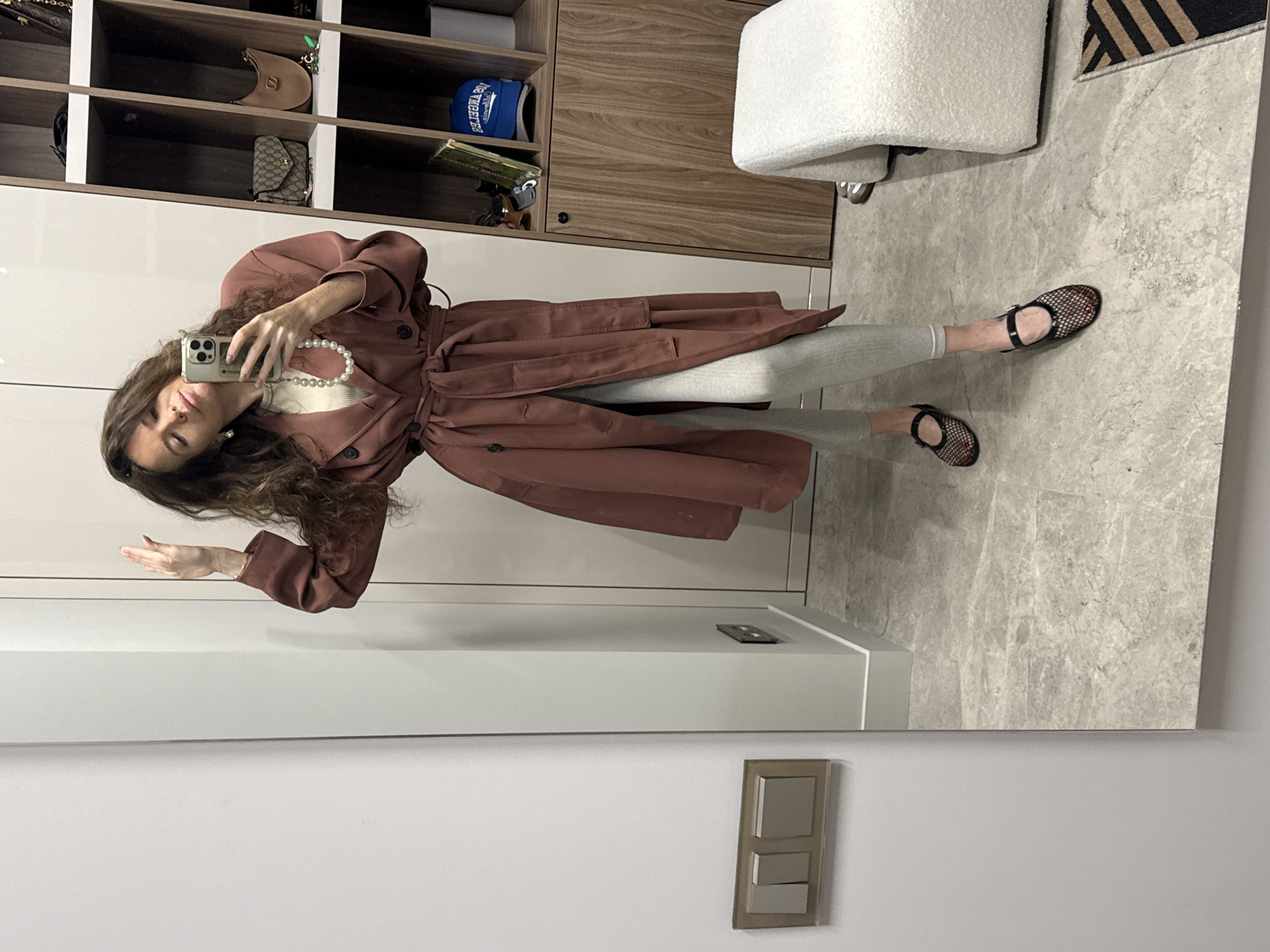 A woman is depicted taking a mirror selfie in what appears to be an indoor setting, likely a dressing room or hallway in Moscow, Russia. The image is a reflection, showing her full body as she holds a silver smartphone with multiple cameras in her right hand to capture the photo.
She has long, wavy dark hair and is wearing a stylish outfit consisting of a reddish-brown, belted trench coat layered over light grey pants. A pearl necklace and matching pearl earrings accessorize her look. Her feet are clad in black mesh-style flat shoes with ankle straps, and her toenails are painted red. She poses with a neutral expression, gazing towards the phone.
The background, visible in the mirror, reveals modern interior design elements. Behind the woman are open shelving units made of light wood with white compartments, holding various items including a brown crescent-shaped handbag, a beige patterned wallet/clutch (featuring a double-G logo), and a blue baseball cap. The text on the cap is visible as a reflection, showing "DETROIT" in reversed letters. To her left, a textured white armchair or stool is partially visible next to a striped black and beige rug. The floor is covered in light grey marble or stone. A white wall section and a gray electrical switch/outlet are also visible at the bottom. The phone screen or case displays small, faint text that might read "firuz" or "finuz".