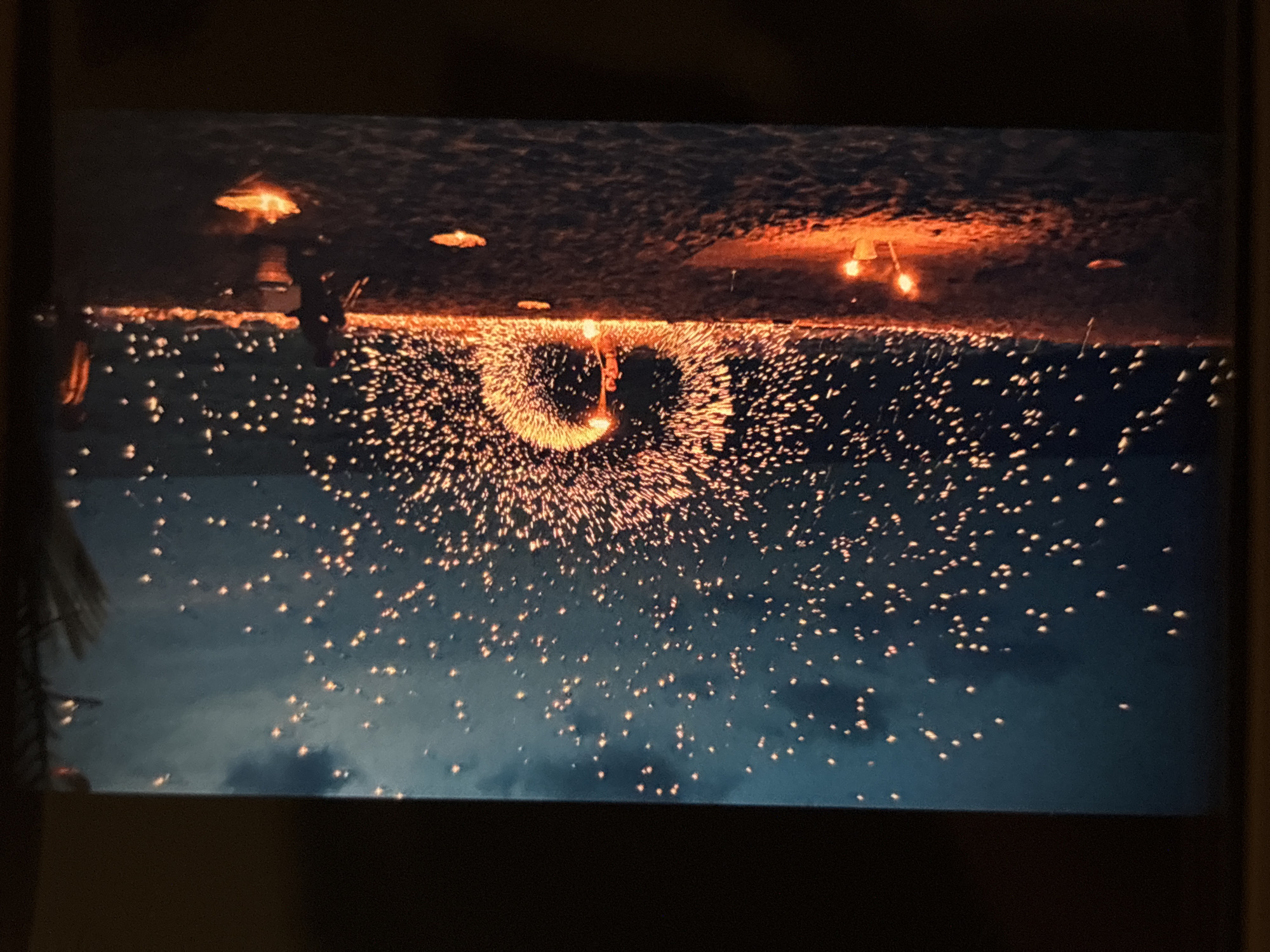 The image depicts a fire performance on a beach in Chalong, Thailand, at dusk. A performer stands in the center, spinning a fiery object that creates a large, circular spray of sparks against the dark, cloud-filled sky. The beach is sandy, with several smaller fires or lanterns scattered on the ground. In the background, the ocean is visible, with gentle waves lapping the shore. The overall atmosphere is dramatic and visually striking, with the warm glow of the fire contrasting against the cool tones of the sky and sea.