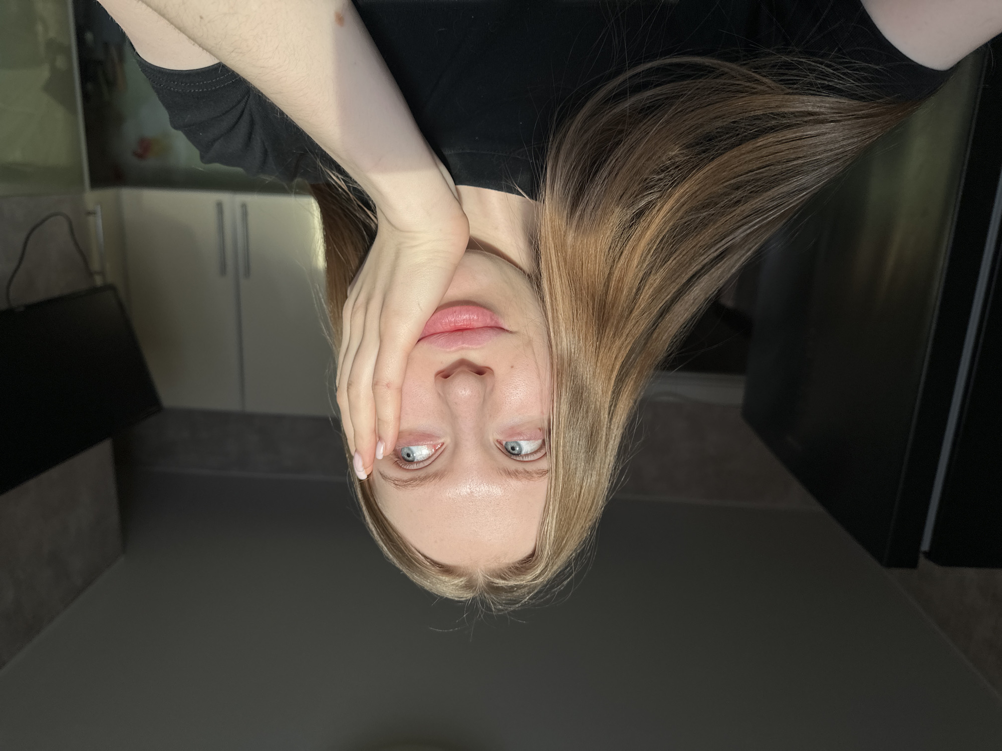 This image depicts a young female in an indoor setting within the city of Chaikovsky, Russia. The subject, a Caucasian female with long, straight, light brown hair and blue eyes, is centrally framed, facing generally towards the camera but with her gaze directed upwards and to her left. She wears a black short-sleeved t-shirt. Her right hand is raised, fingers slightly splayed, resting against her right cheek and temple, with her lips slightly parted. Strong, direct sunlight illuminates the right side of her face, hair, and arm, creating a distinct shadow line across the scene. The background suggests a residential kitchen environment. To the left, a dark, vertical appliance, likely a refrigerator, is partially visible. To the right, light-colored upper kitchen cabinets with two vertical metallic handles are present, beneath which a dark, wall-mounted television or monitor with visible cables is installed. Elements of a countertop and backsplash are discernible below the television.