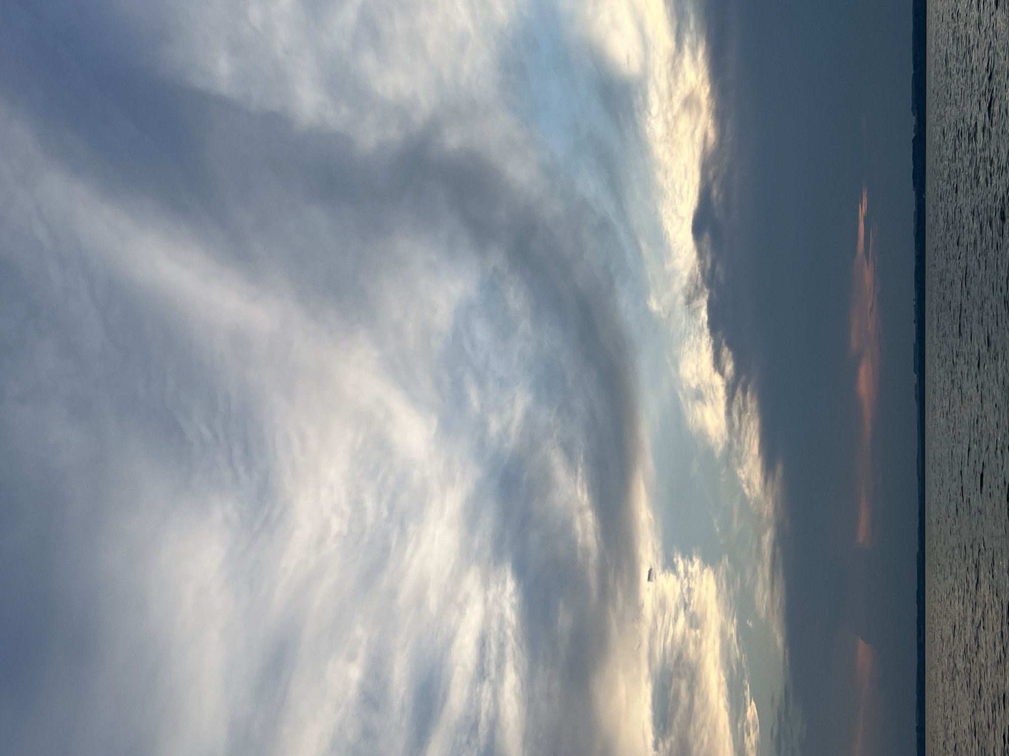 The image depicts a body of water, likely a lake or a wide river, under a dramatic sky filled with clouds. The clouds are varied in texture and color, ranging from wispy white streaks to darker, more dense formations. Some areas of the sky show hints of golden light, suggesting either sunrise or sunset. A distant, dark shoreline with faint indications of buildings or trees is visible along the horizon. The water's surface is rippled, reflecting the muted colors of the sky. There are no people or animals visible in the scene. The overall atmosphere is calm and natural. The location is identified as Novopichugovo, Russia.