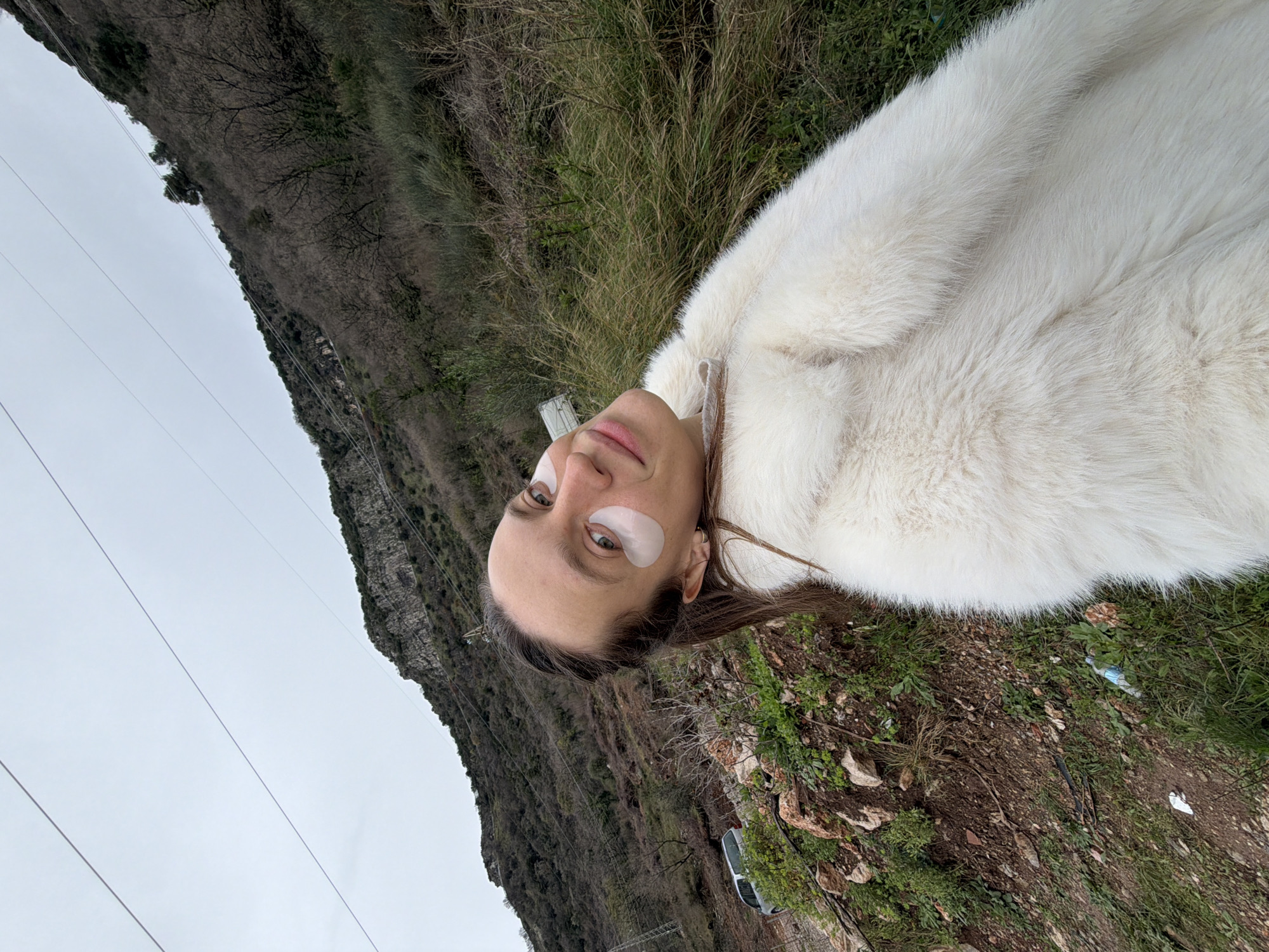 The image displays a female subject in a selfie-style portrait, positioned outdoors on a hillside. The subject has light brown hair, pulled back, and wears two white under-eye patches, one beneath each eye, along with a white faux-fur coat, exhibiting a subtle smile. The immediate foreground features exposed reddish-brown dirt, rocks, and sparse green and dry vegetation. In the mid-ground to the subject's left, a silver-colored SUV is partially visible parked on what appears to be a dirt track or shoulder. The background is dominated by a large, vegetated mountain under an overcast grey sky. Multiple power lines are visible extending diagonally across the upper portion of the frame. Small pieces of litter, including a blue plastic bottle and a white wrapper, are present on the ground in the lower right. This scene is located in a mountainous, natural area within the Budva municipality of Montenegro, likely along a rural road or trail on its periphery.