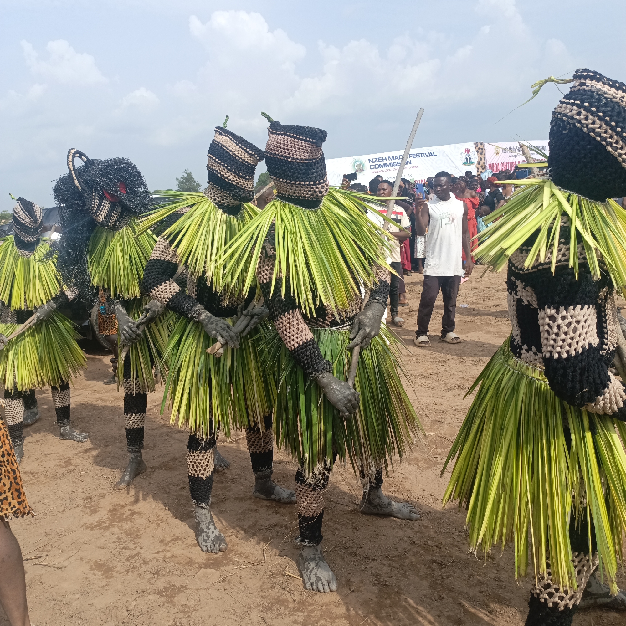 The image captures a vibrant cultural event in Akwanga, Nigeria. A group of individuals are adorned in traditional masquerade costumes, featuring black and beige woven headpieces and body coverings, complemented by skirts of green palm fronds. They are standing on a dirt ground, with mud caked on their bare feet and hands.
In the background, a crowd of spectators is visible, some holding mobile phones, likely capturing the spectacle. A banner or sign can be seen in the distance, suggesting a festival or organized event. The sky above is partly cloudy, providing ample daylight for the outdoor setting. The overall atmosphere is one of cultural celebration and community engagement.