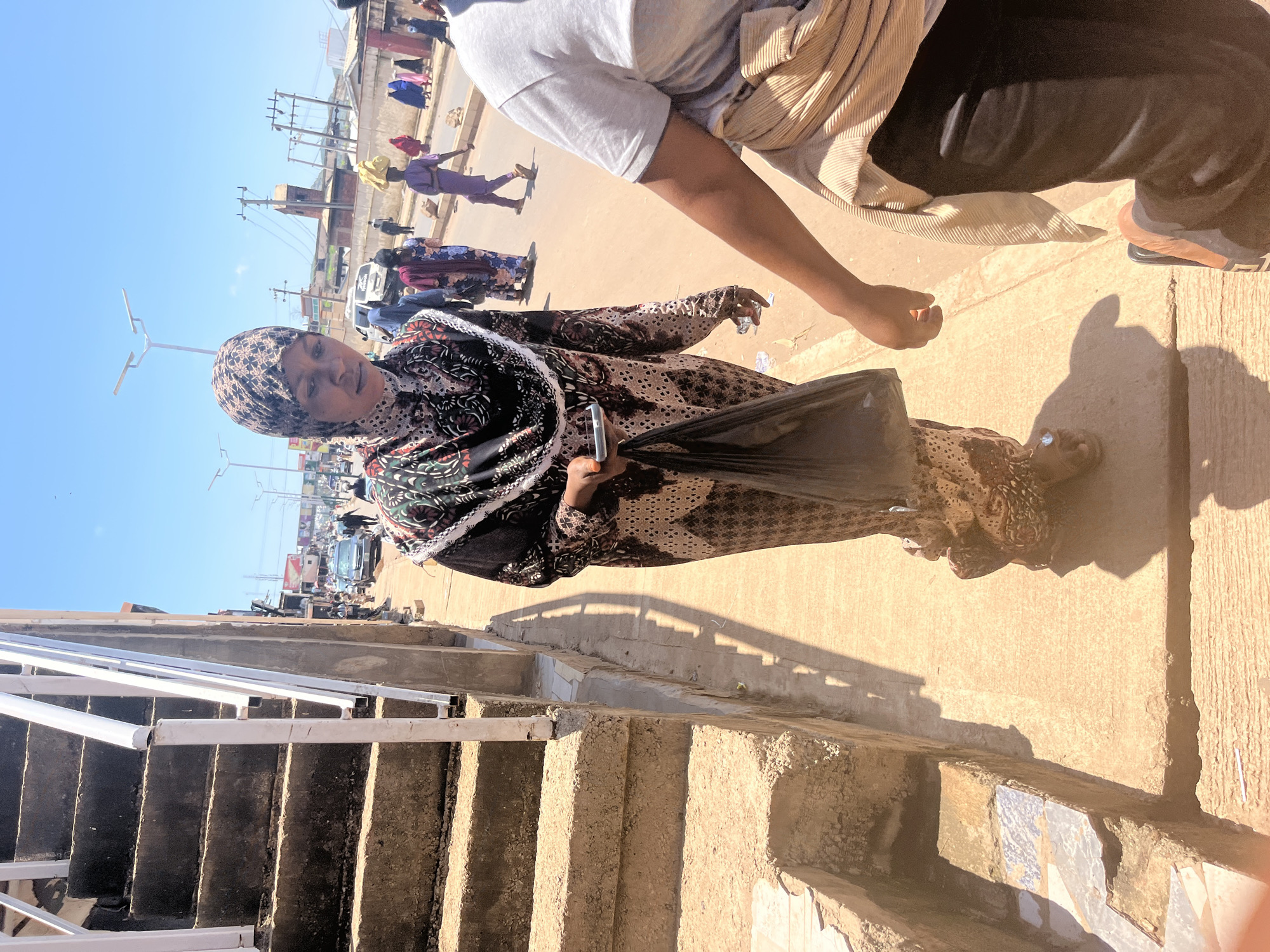 In Jos, Nigeria, a woman wearing a patterned hijab and a long, flowing dress walks on a sidewalk next to a concrete staircase with white railings. She is holding a black plastic bag in her left hand and a mobile phone in her right. The sidewalk is made of concrete and shows signs of wear. Behind her, people are walking on the street, some carrying items on their heads. Cars are visible on the road, and there are buildings and utility poles in the background. The sky is clear and blue, indicating a sunny day. The scene appears to be a busy urban area.