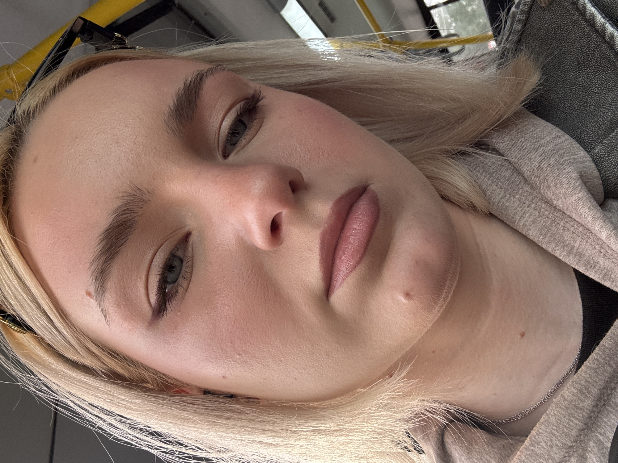 A blonde woman with light skin, wearing makeup, is looking down. She has on a grey hoodie over a black shirt, with a silver chain necklace. Her hair is blonde and shoulder-length. The background appears to be a public transportation vehicle, possibly a bus, with yellow handrails visible. The scene is set within the city of Sinyavino, Russia.