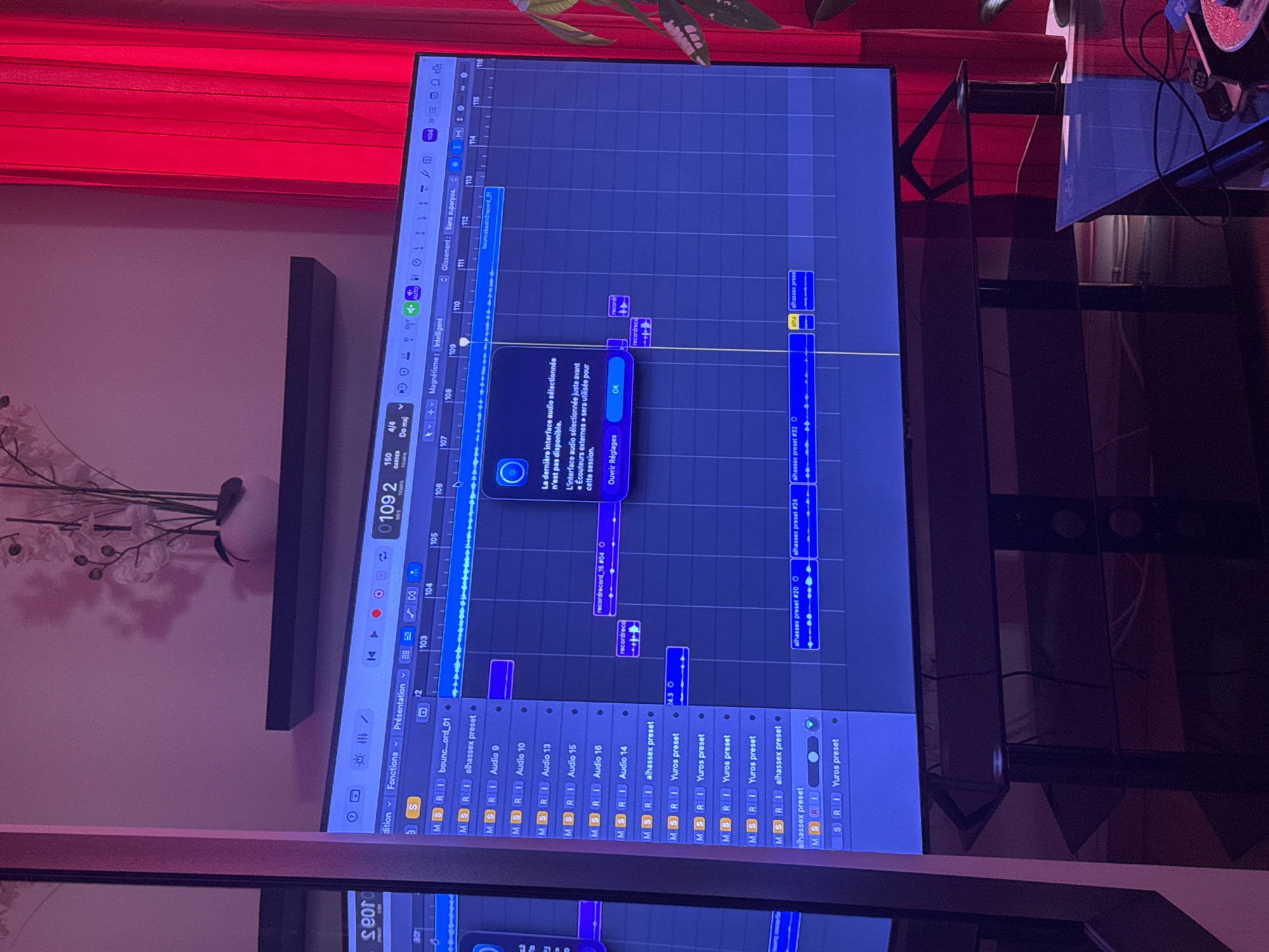 The image shows a screen displaying audio editing software, likely Ableton Live, running on a computer or monitor. The screen shows a session view with multiple audio tracks, including tracks named "bounc...ord_01", "alhassex preset", "Audio 9", "Audio 10", "Audio 13", "Audio 15", "Audio 16", and "Audio 14", along with tracks named "Yuros preset" and "alhassex preset". Audio clips are arranged across the tracks, forming a musical arrangement. A popup message is visible on the screen in French, stating that the last selected audio interface is not available and that the "external headphones" interface will be used for the session. The software interface includes transport controls (play, stop, record), a tempo display (109.2 BPM), and arrangement navigation tools. The environment surrounding the screen is dimly lit. A red curtain is visible in the upper left, and a blurred image of an object that may be a plant is located above the monitor. There's a shelf or ledge visible behind the monitor. The context suggests an indoor studio or home environment. There is no indication that the photo was taken in Paris, France.