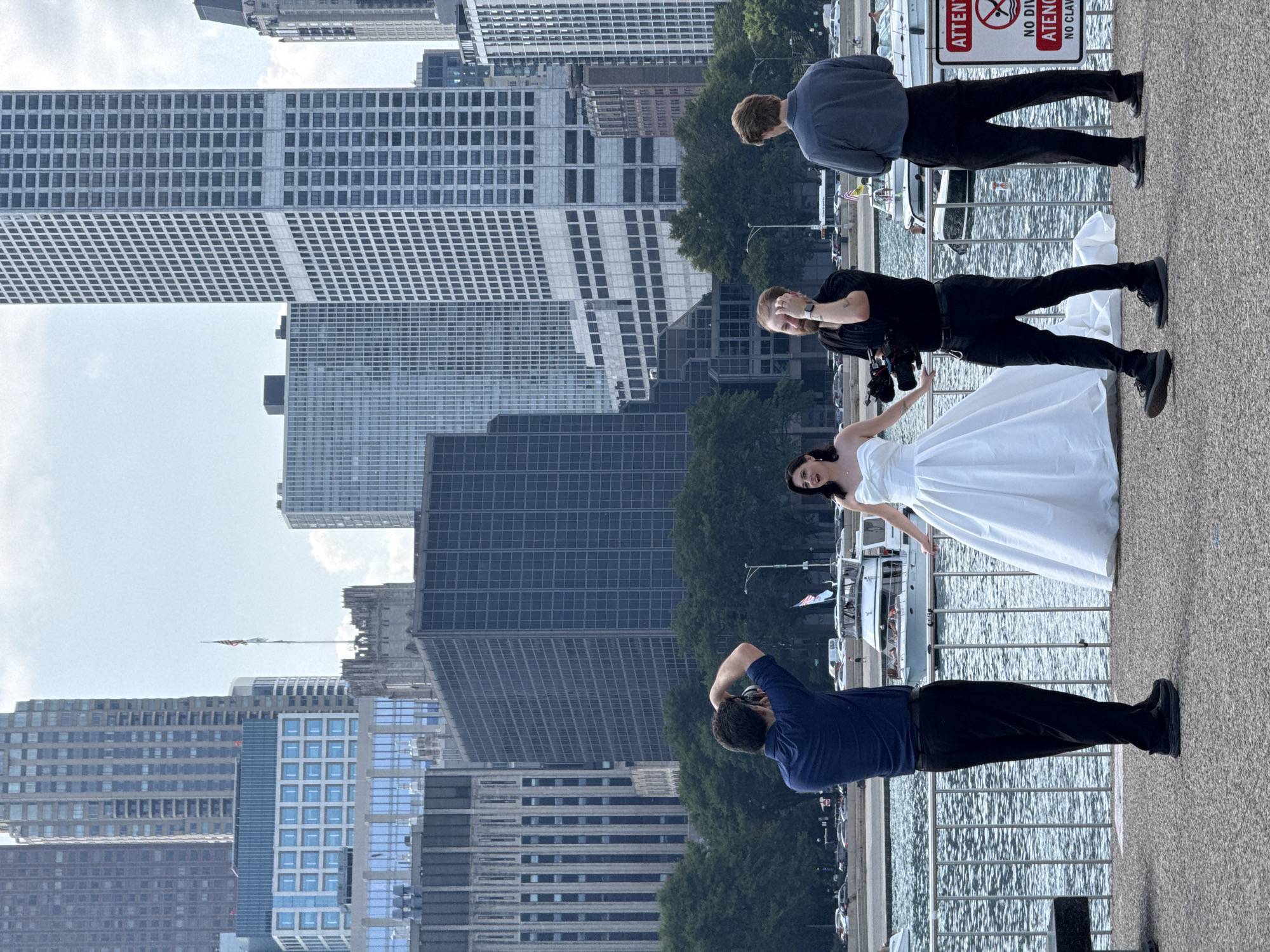 The image captures a wedding photoshoot with the Chicago skyline as a backdrop. In the foreground, a bride in a white gown stands by a railing, facing right and smiling. To her left, a photographer in a navy blue shirt is taking pictures with a DSLR camera. To her right, a videographer in a black shirt is operating a video camera, and another person in a gray shirt stands behind him. The group is positioned on a paved area overlooking a body of water, likely Lake Michigan, with several boats visible. Behind them, the distinctive architecture of Chicago's high-rise buildings rises against a cloudy sky. A sign with red lettering, partially obscured, is visible on the right side of the image.