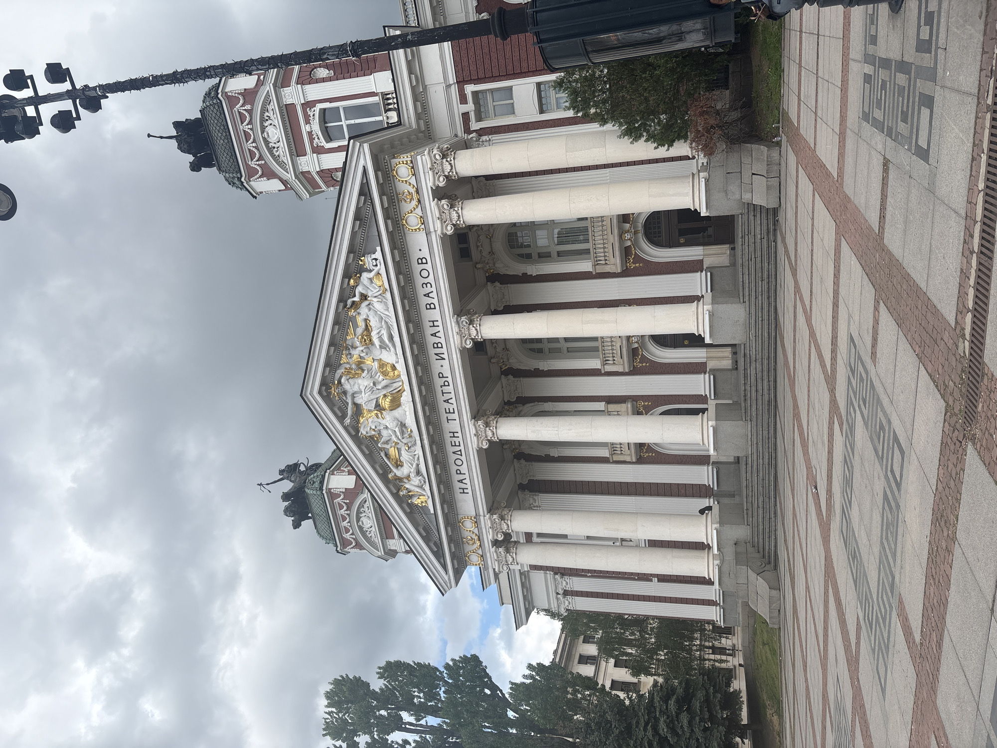 The image shows the Ivan Vazov National Theatre in Sofia, Bulgaria. The architecture is neoclassical with white columns and a triangular pediment decorated with sculptures. The building is red and white with ornate details. In the foreground, there is a paved square with geometric patterns. The sky is overcast.