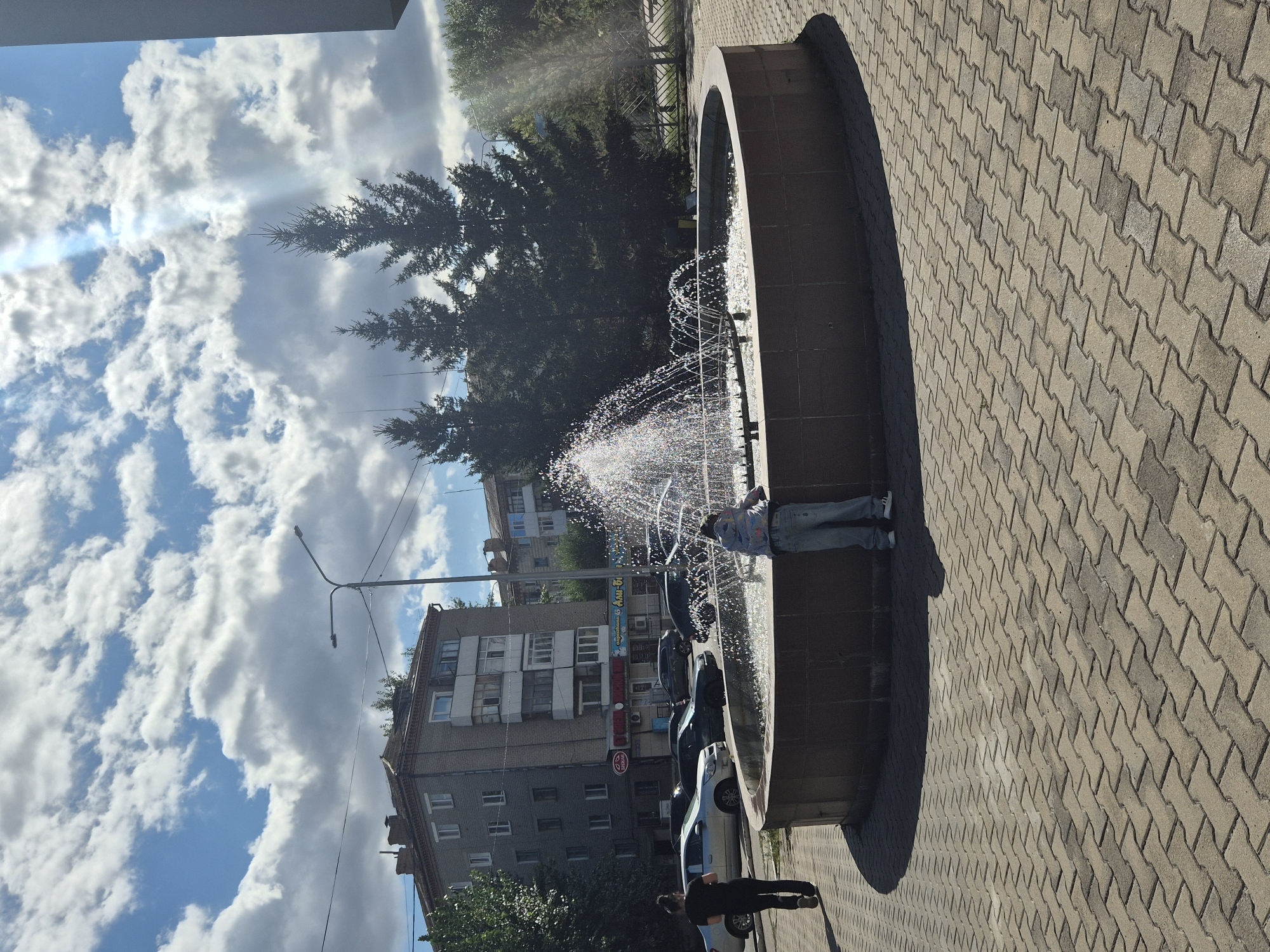 In Kokshetau, Kazakhstan, a person stands near a circular fountain on a paved area. The fountain is active, with water spraying upwards. The person is wearing blue jeans and a hooded jacket. Another person in dark clothing and pants walks away from the fountain. Buildings, including a multi-story brick structure with balconies, are visible in the background. Several cars are parked on the street. Evergreen trees are present behind the fountain. The sky is blue with scattered white clouds.