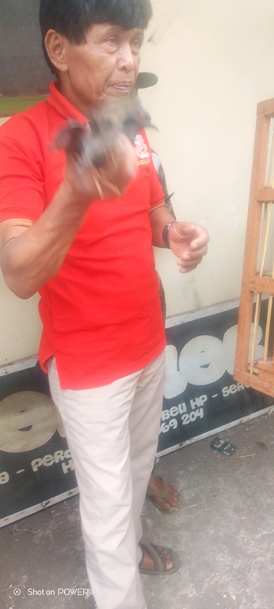 An older man with short black hair, wearing a red polo shirt and light-colored pants, stands outdoors. He is holding a small bird in his right hand, which is extended towards the camera. The man's left hand is also visible, and he appears to be looking down at the bird. In the background, there is a light-colored wall and a wooden birdcage. A portion of a dark sign with white lettering is visible on the ground in front of the man, with a small mobile phone lying on it. The ground appears to be concrete or asphalt.