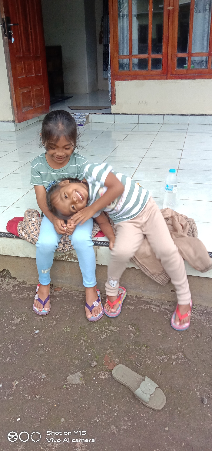 This image depicts an outdoor scene in Ranugedang, Indonesia, featuring two young females. An older girl, wearing a light green striped short-sleeved shirt and light blue leggings, is seated on a raised concrete ledge. She has dark hair and a smiling expression. The younger girl is positioned horizontally across the older girl's lap, with her head tilted back and a wide smile on her face. She wears a light green and white striped short-sleeved shirt, light brown leggings, and bright red and pink flip-flops. The older girl wears purple and pink flip-flops. A red and brown plaid fabric item is placed beneath the older girl on the ledge. To the right of the subjects, a clear plastic water bottle with a blue cap rests on a white tiled surface. The foreground consists of uneven, dark brown dirt ground with small stones and sparse dry vegetation. A single light green open-toed sandal lies on its side on the dirt. Behind the subjects is a building with a light-colored tiled porch floor, a cream-colored exterior wall, and a partially open brown wooden door on the left, revealing a darker interior. To the right of the door is a brown wooden-framed window with multiple panes, reflecting ambient light.