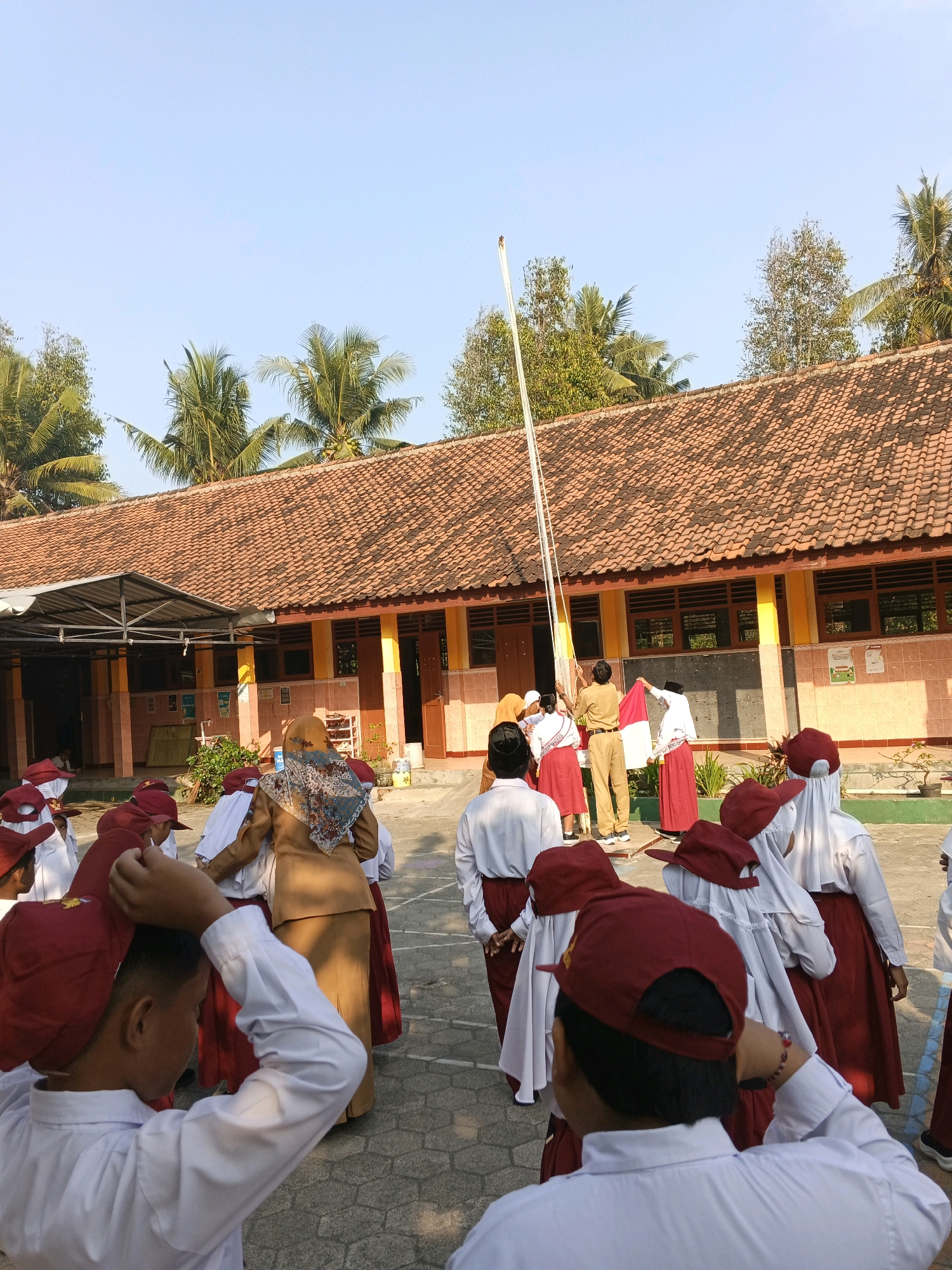 In Glagah, Indonesia, at a school, a group of schoolchildren in white shirts and red skirts or shorts, many wearing matching red hats, are gathered in a courtyard. Several children are seen from behind, adjusting their hats. In the background, a teacher in a brown uniform and a woman in a brown headscarf and patterned hijab are interacting with other students near a flagpole. One student holds a red and white flag, suggesting a flag-raising ceremony. The school building has orange tiled roofs and yellow walls, with visible windows and doors. Palm trees are visible above the building, indicating a tropical climate. The ground is paved with interlocking stones. The sky is clear and blue, with the sun casting shadows, suggesting it is daytime.