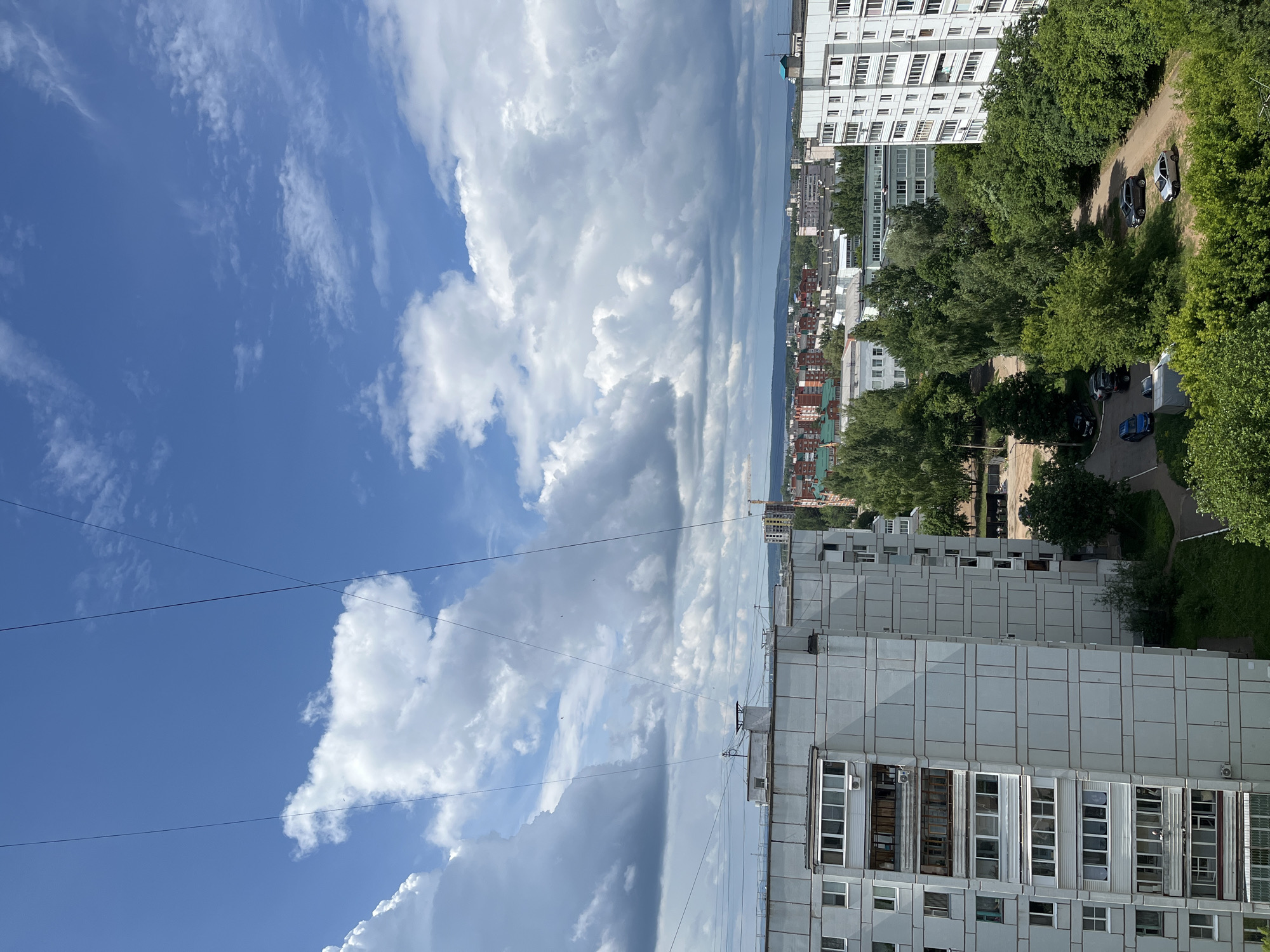 An aerial view captures a residential area in the city of Chaikovsky, Russia, under a bright blue sky dotted with fluffy white clouds. In the foreground, the side of a large, light-grey apartment building is visible, featuring numerous windows and balconies. Beyond this building, more apartment complexes and other structures stretch into the middle distance, interspersed with abundant green trees. A construction crane is visible in the far distance on the right side of the image, indicating ongoing development. The scene is peaceful and appears to be a typical day in a Russian urban environment.