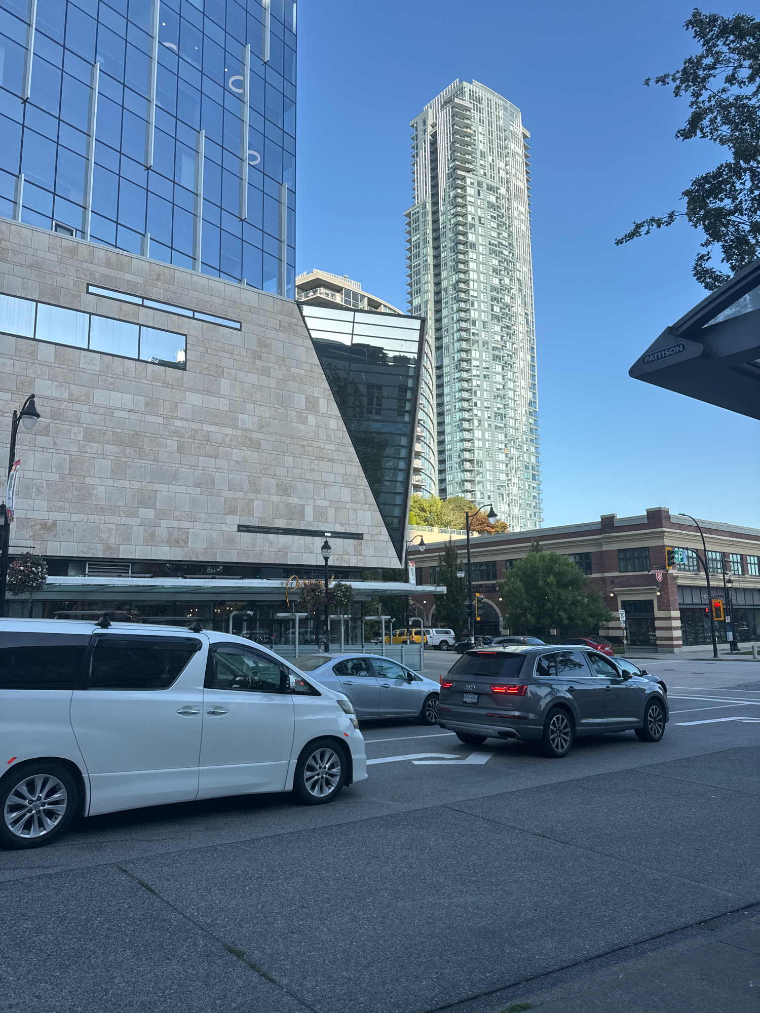 In New Westminster, Canada, a street-level view captures a modern urban scene under a clear blue sky. In the foreground, a white minivan and a grey SUV are driving on a paved road. To the left, a contemporary building with a stone facade and large glass windows stands tall. Further in the background, a very tall, slender skyscraper dominates the skyline. Across the street, a red brick building with visible storefronts and architectural details is situated. A bus shelter with "PATTISON" signage is partially visible in the upper right corner. The environment appears to be a commercial or downtown area, indicated by the buildings and traffic.