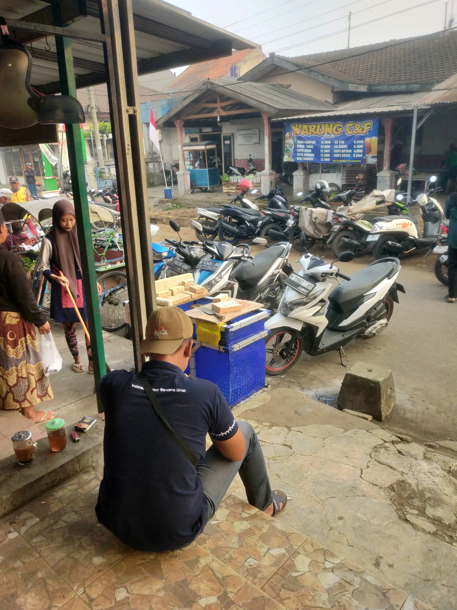 The image depicts a street scene in Bululawang, Indonesia, likely in front of a small business or food stall. In the foreground, a person wearing a dark blue t-shirt and grey pants sits on a tiled step, facing away from the camera. The t-shirt has white lettering on the back that reads "Indonesia Bersama Umat". Next to the person are two glasses, one with a dark liquid and the other with a lighter colored liquid, and a small brown object that appears to be a book or small box, and a cigarette lighter. The background shows a street lined with parked motorcycles. A banner displaying food and drink items with prices is visible on the wall of a building. Several people are seen in the background, including a woman in a brown headscarf and a patterned skirt standing near a cart, and other individuals walking or standing. The architecture suggests a casual, possibly rural or suburban, commercial area. The overall atmosphere is one of everyday street life.