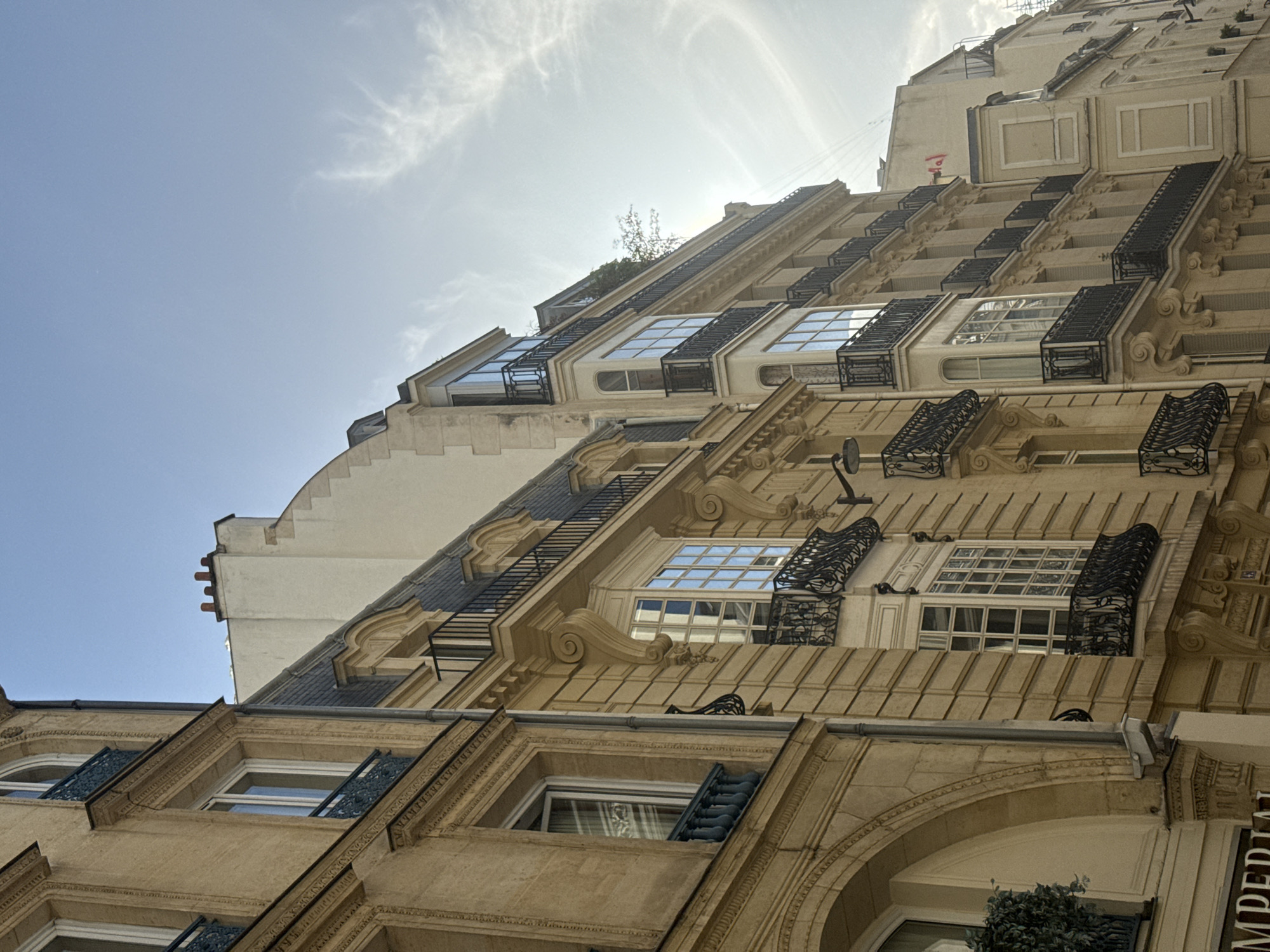 This low-angle photograph captures the ornate facade of a multi-story building, likely an apartment block or hotel, under a bright, sunny sky. The building, characteristic of Parisian architecture, is constructed from light-colored stone or stucco and features numerous windows, many adorned with intricate black wrought iron balconettes and railings. Decorative moldings, cornices, and scrollwork embellish the facade. Higher up, dormer windows are integrated into a dark mansard roof. A clear blue sky with scattered wispy clouds occupies the upper left, with the sun's glare visible towards the top right. A small amount of green foliage can be seen on an upper-level balcony and in a lower window box. A satellite dish is attached to the facade. Part of the word "IMPERIAL" is visible vertically on the right side of the building. The time of day is bright daylight, and no people or specific activities are depicted. The overall impression is one of classic Parisian urban elegance.