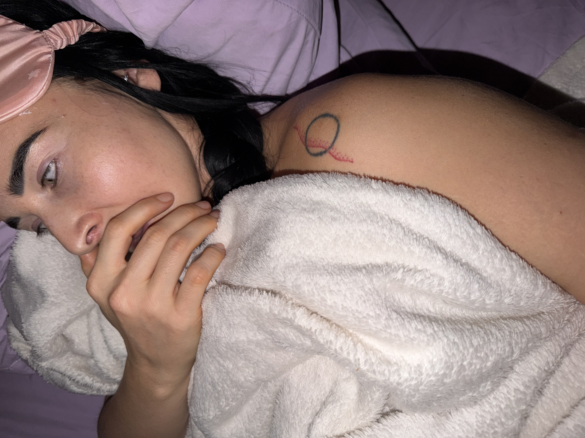 The image shows a close-up of a person, likely female, lying down in what appears to be a bedroom. Her dark hair rests on a light purple pillow or sheet, and a pink satin sleep mask is pushed up onto her forehead. She is partially covered by a thick, white, fluffy blanket or towel. Her left hand is placed near her mouth and chin, partially obscuring her lower face. Her eyes are partially open and gaze upwards and to the left, suggesting a state of rest, contemplation, or waking.
A notable detail is a tattoo on her right shoulder: a black circular outline with a red, abstract line or 'tail' extending from it, possibly resembling a stylized 'Q'. The lighting in the scene is dim, implying it could be early morning, late night, or a dimly lit indoor space. The overall atmosphere conveys a sense of quietude or repose. No specific location cues are visible within the image, but the provided context places the location as Unknown, Russia. There is no visible text.