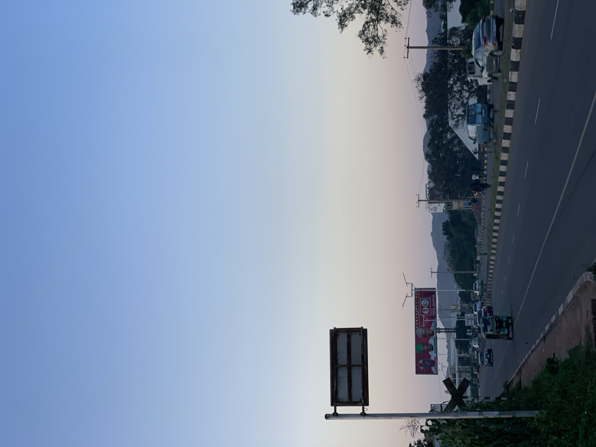 The image depicts a view of a road in Jos, Nigeria, at dusk. A highway is visible with multiple lanes, bordered by a concrete median and a grassy embankment. Several vehicles are on the road, including cars, a tricycle, and a truck. A large billboard featuring three men in traditional attire is prominently displayed on the left side of the road, with text that reads "HUMANITY FIRST" and "FINAL BATTLE." A street sign or traffic signal is also present on a pole. In the background, trees and hills are silhouetted against a gradient sky transitioning from blue to pale pink, indicating the time of day. The environment appears to be an urban or semi-urban area, with signs of ongoing activity and infrastructure.
