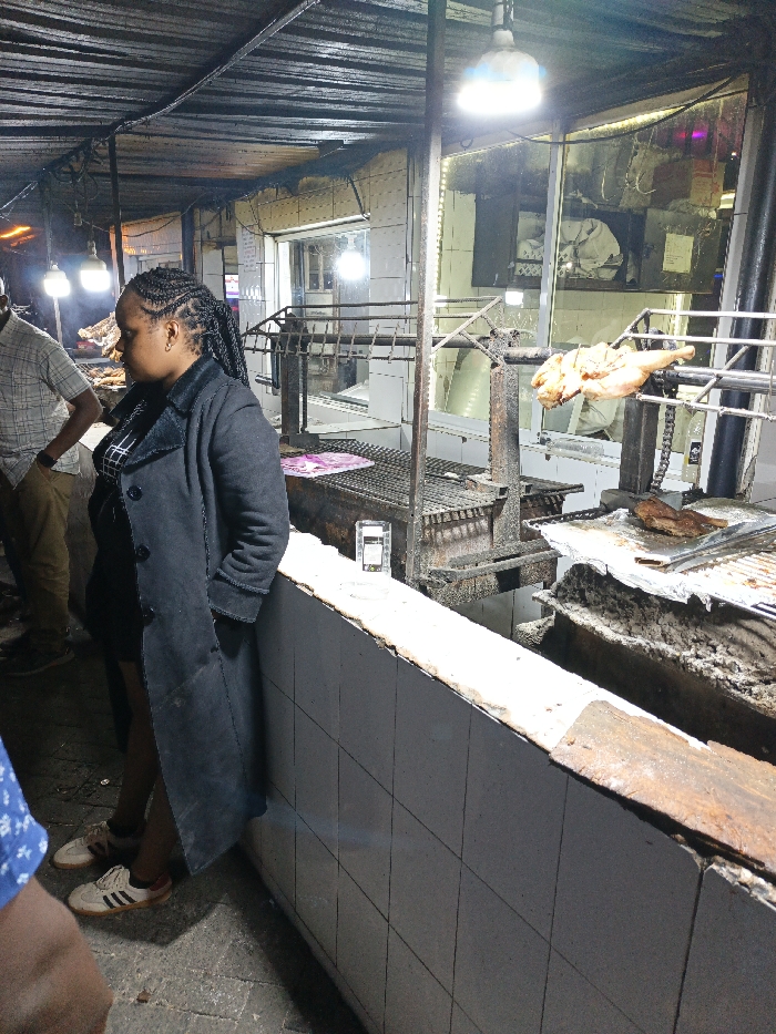 The image displays a nighttime scene within an open-air or semi-enclosed food establishment, typical of a nyama choma (grilled meat) restaurant in Nairobi, Kenya.
In the foreground-left, a female individual with dark braided hair stands, facing towards the right. She wears a dark, possibly black or dark grey, long coat over a checkered top and white sneakers with dark stripes. Her hands are in her pockets. To her immediate left, part of a male individual wearing a plaid shirt and tan pants is visible.
The central and right portions of the frame are dominated by a large, active grilling station. A whole, partially cooked chicken is mounted on a mechanical rotisserie spit, positioned directly over a charcoal grill. To its right, another section of the grill holds pieces of meat, possibly beef or goat, resting on a foil-covered tray above hot coals. The metal grilling equipment is robust and shows signs of heavy use. A small, transparent, rectangular object, resembling a smartphone or a small digital display, is placed on the low, white-tiled partition that separates the customer area from the cooking space.
Behind the grilling area, a brightly lit building interior is visible through large glass windows. The interior features white-tiled walls and appears to be a commercial kitchen or food preparation zone, with a blurred figure, likely staff, discernible inside. Overhead, a dark, corrugated metal roof is supported by metal poles, from which several bright, exposed light bulbs are suspended, illuminating the scene. The ground beneath the individuals is a dark, paved surface. The overall environment suggests a casual, bustling eatery focused on grilled meats, a common feature in Nairobi's culinary landscape.