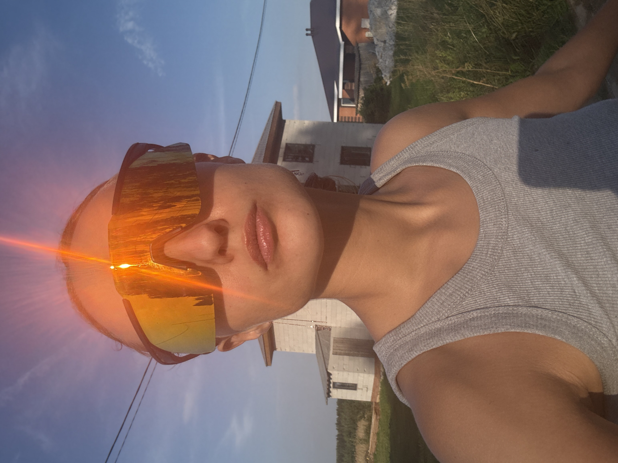 This is a close-up, vertically oriented, selfie-style photograph taken outdoors. The primary subject is a person, whose face is partially visible wearing large, reflective sunglasses that mirror a sunset over water. The person's skin appears tanned, and they are wearing a grey ribbed tank top. The background features a bright blue sky with some wispy clouds, indicating it is likely daytime.
In the middle ground, there are unfinished buildings, suggesting a construction site or a rural area under development. The vegetation in the background appears dry and grassy, possibly indicative of a summer or early autumn season. Power lines are also visible, crossing the sky.
A prominent lens flare, appearing as a bright orange and yellow streak with radiating lines, emanates from the left side of the image, partially covering the sunglasses and the subject's face. This lens flare, along with the sunset reflection in the sunglasses, suggests the photograph was taken during the golden hour, shortly before or after sunset. The overall mood conveyed is relaxed and perhaps contemplative, given the subject's partial view and the scenic, yet somewhat undeveloped, setting. The location is identified as Podshivalovo, Russia. There is no visible text in the image.