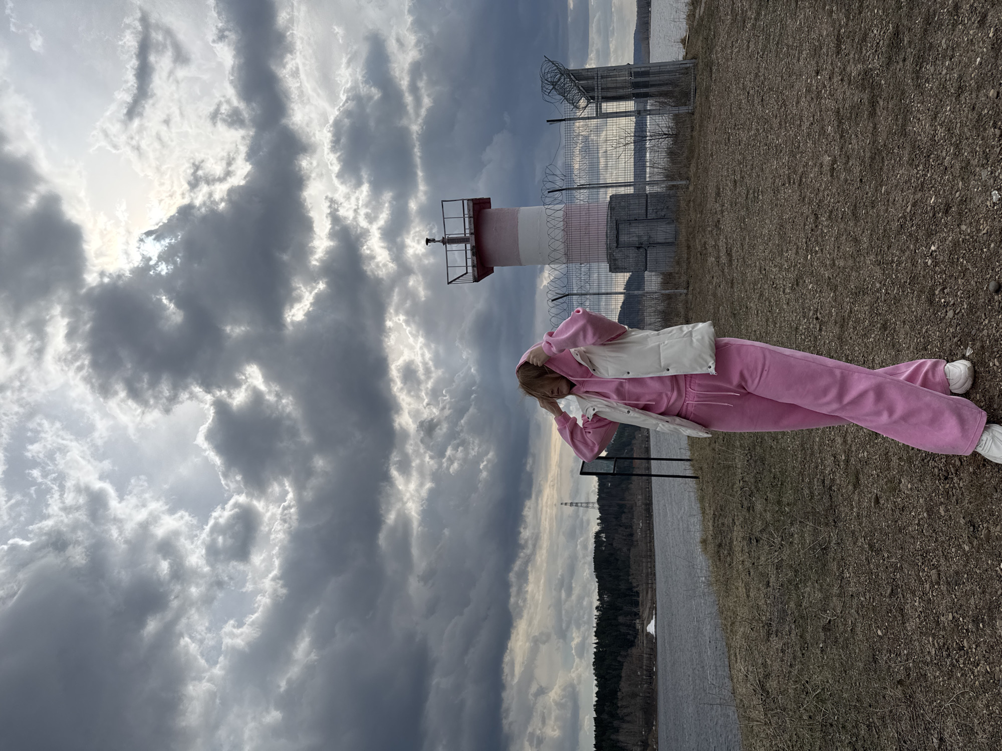 An outdoor, eye-level shot captures a person standing on a gravelly and dry grassy bank beside a large body of water. The subject, positioned slightly to the left of the frame's center, wears a bright pink tracksuit with a white puffy vest and white sneakers. Their head is tilted, and one arm is raised towards their head, partially obscuring their face.
Behind the person, towards the right, stands a distinctive red and white striped structure resembling a lighthouse or a communication beacon, surrounded by a metal fence topped with barbed and razor wire. Beyond the fence, the expansive body of water extends into the background, with a distant treeline and another slender transmission tower visible on the far shore.
The sky is heavily overcast with dramatic, dark grey clouds, though patches of bright light suggest the sun is breaking through or behind the clouds, creating illuminated edges. The overall weather appears cool and cloudy. The scene depicts a rugged, natural landscape combined with industrial or navigational elements. No visible text is present in the image. The location context is Chaikovsky, Russia.