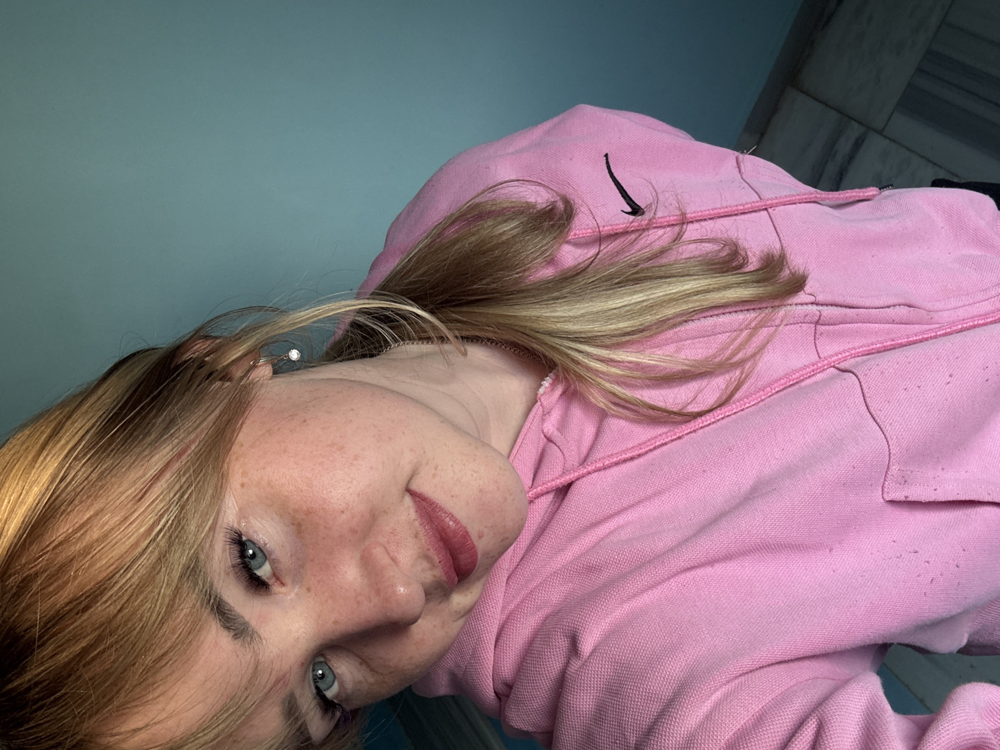A close-up, angled selfie captures a young woman, likely in Марковский, Russia, with her head tilted to one side. She has fair skin, prominent freckles across her nose and cheeks, and bright blue eyes with defined eyelashes. Her blonde hair is parted to the side and cascades over her shoulders. She wears a soft, light pink lipstick, and her expression is a gentle, engaging smile directed towards the camera.
She is dressed in a light pink hooded sweatshirt (hoodie) which features a distinct black Nike swoosh logo on the upper left chest area. The hoodie's fabric shows some signs of wear, such as pilling. Pink drawstrings for the hood are visible around her neck. The background consists of a solid teal-blue wall on the left side of the frame and a darker, possibly tiled or patterned wall on the upper right, suggesting an indoor setting. The lighting is ambient, indicative of an interior space. The woman appears to be taking a self-portrait, engaging directly with the viewer.