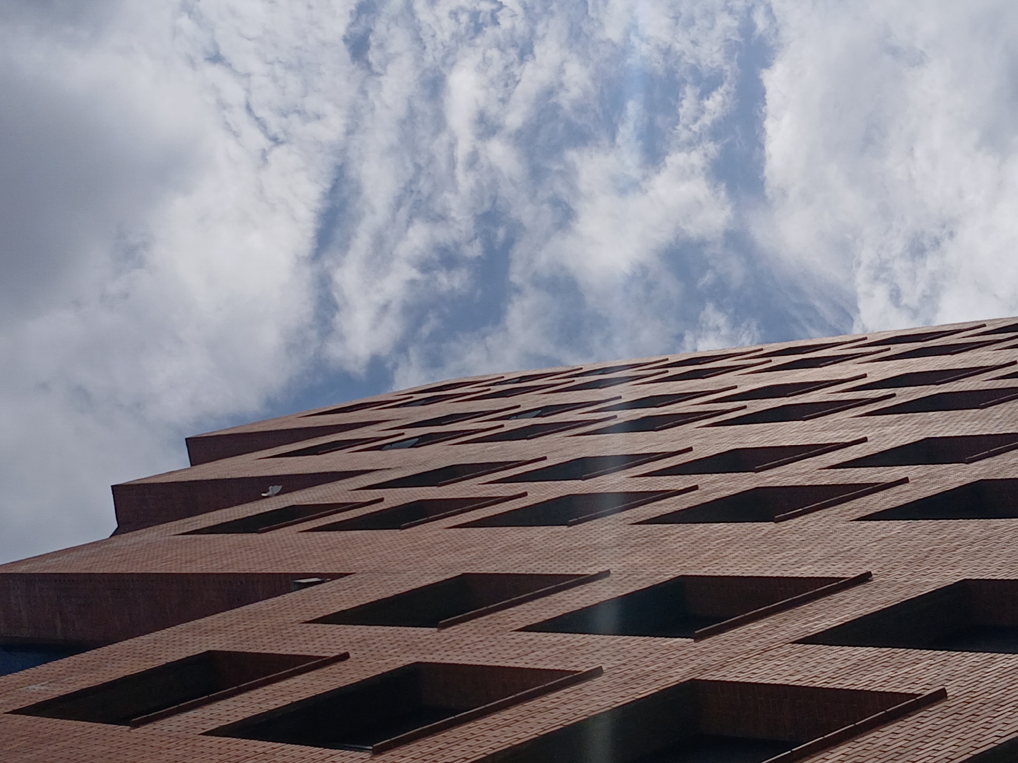 The image presents a low-angle shot of a multi-story building made of reddish-brown brick. The facade features numerous rectangular window openings, evenly spaced in a grid-like pattern. The perspective is upward, revealing the textured brick surface and the geometric arrangement of the windows. Above the building, a cloudy sky fills the upper portion of the frame. The clouds are white and streaky, contrasted against a light blue sky. The location in the city of Ciudad Guayana, Venezuela, is not readily apparent within the visual elements of the photograph.