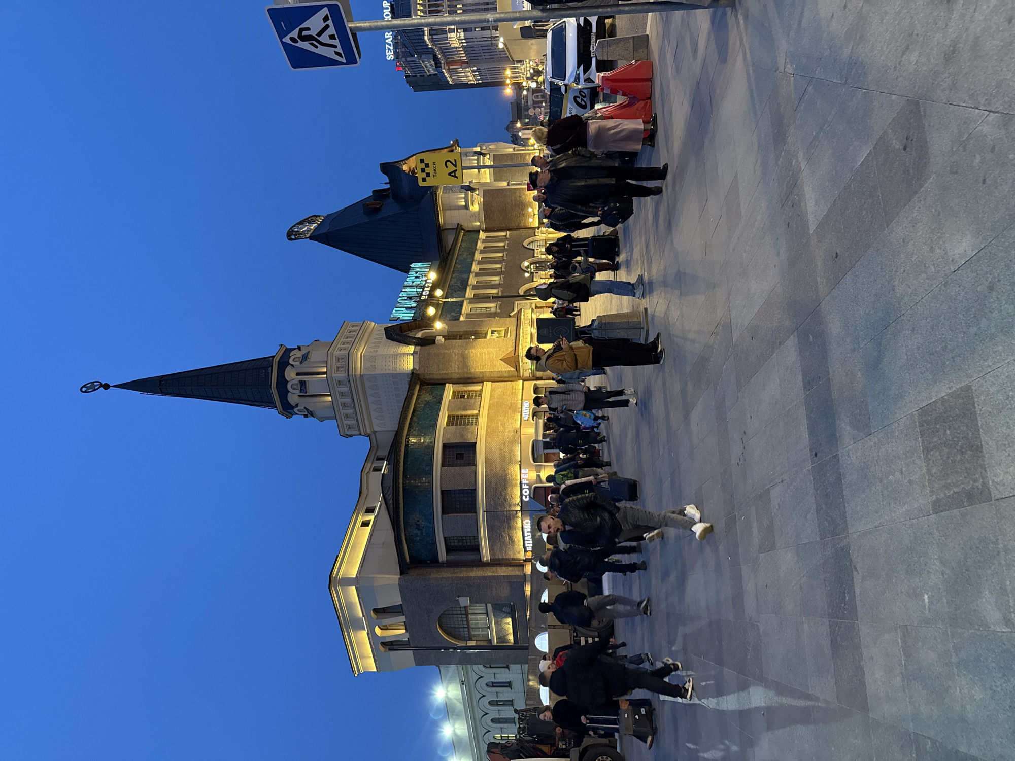 This urban scene in Moscow, Russia, captures a bustling square during the blue hour, suggesting either early morning or evening. The sky is a deep, clear blue, contrasting with the warm artificial lights illuminating the buildings and pavement.
Numerous people are visible across the foreground and midground, many with luggage, indicating a travel hub or station environment. They are dressed in coats and casual wear, standing, walking, and conversing. The ground is paved with large, grey stone tiles, reflecting light and creating elongated shadows from the people and structures.
To the left, a grand building with a distinctive spired tower and classical architectural elements stands prominently, its details highlighted by uplighting. Further right, a more modern building is also illuminated, featuring visible text such as "COFFEE" and "В ПУТЬ!" (meaning "On the way!" or "Bon Voyage!"). In the background, tall city buildings are visible, with "SEZAR GROUP" text on one.
Other notable details include a blue pedestrian crossing sign in the upper left, a yellow "TAXI" sign with "A2" indicating a designated pick-up area, and a white car with a "Go" logo partially visible near red traffic barriers. The overall impression is one of a busy, active city center, likely a transportation interchange, with people on the move.