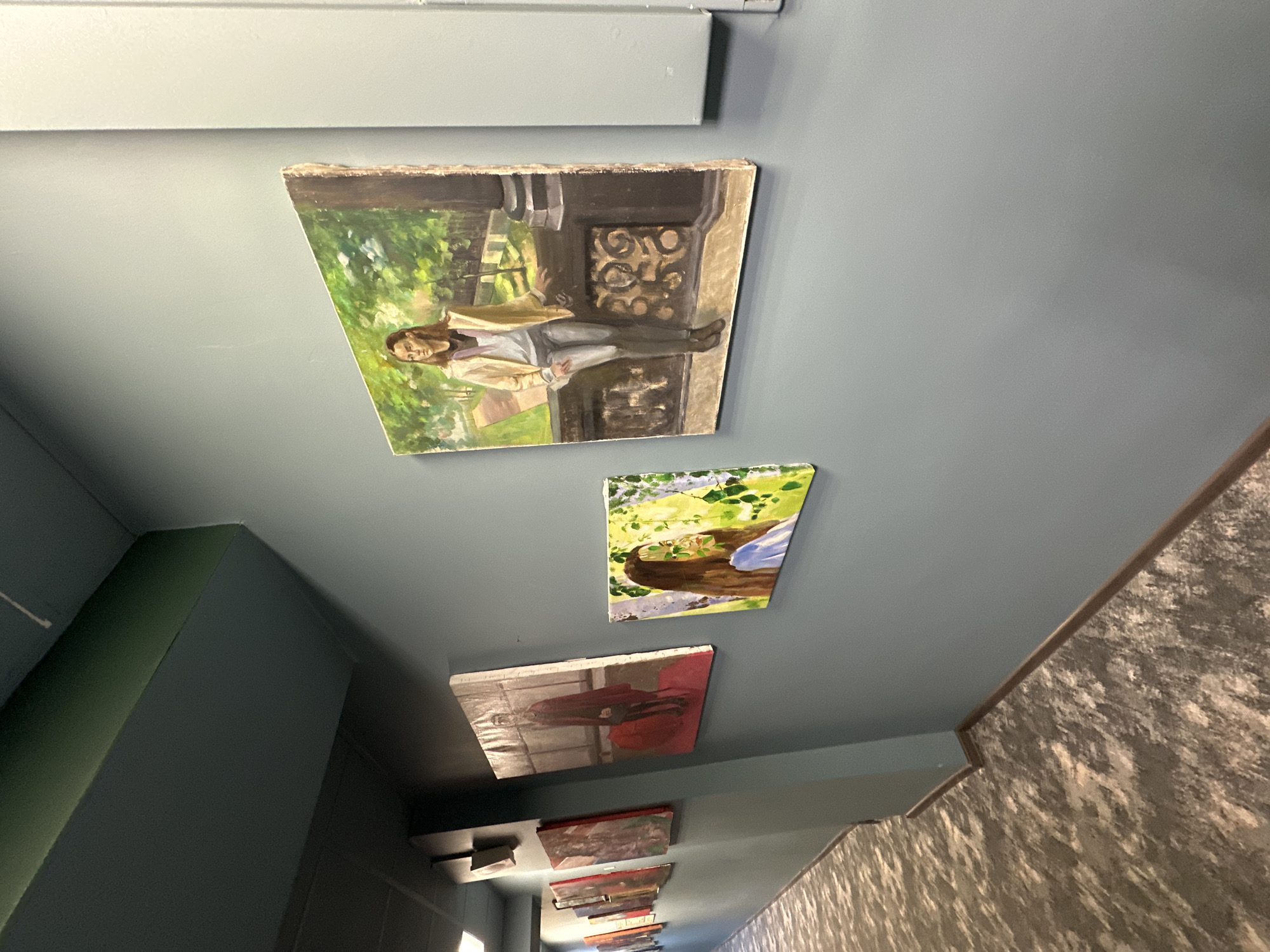 The image shows a hallway ceiling in Saint Petersburg, Russia. A series of small paintings are affixed to the ceiling. The top painting depicts a person sitting on a wrought iron structure in a green outdoor setting. Below are two other paintings of portraits. Several more paintings extend into the distance, growing smaller and less distinct. The hallway walls are painted in gray and green hues with a decorative textured wall visible on one side.