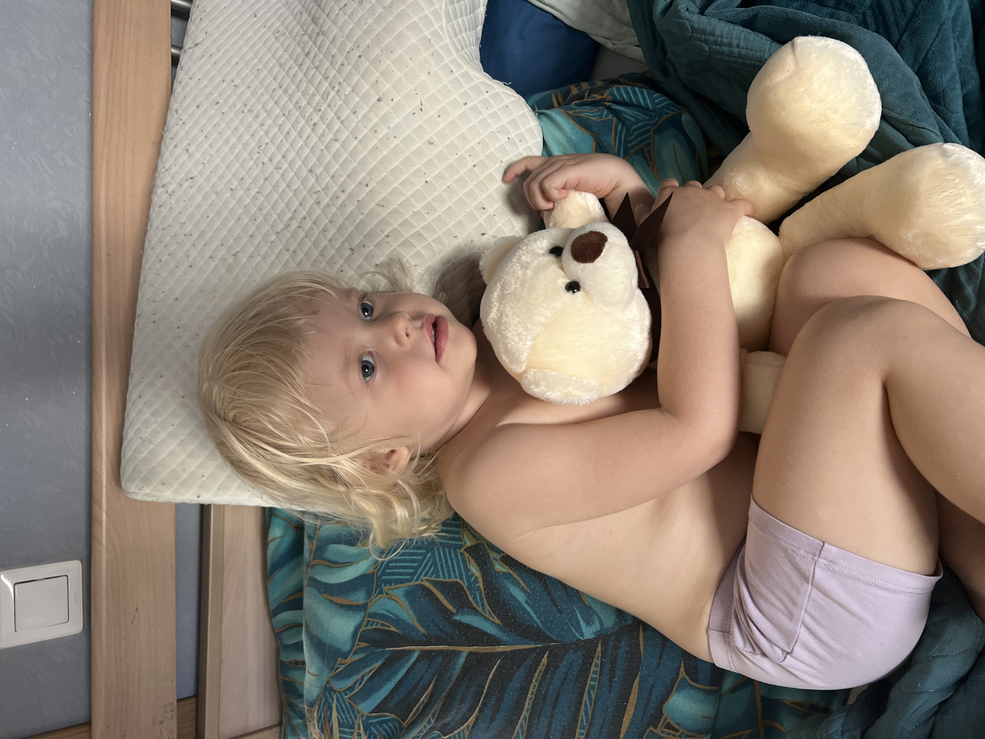 This indoor image features a young child, likely a toddler, with blonde hair and blue eyes, lying on their back on a bed. The child is shirtless and wearing light purple underwear or shorts, looking directly at the camera with a calm, slightly open-mouthed expression.
Cuddled against the child's chest is a light cream-colored plush toy, resembling a bear or lion, with a brown snout and dark eyes, and what appears to be a brown mane around its head. The child's arms are wrapped around the toy.
The bed is adorned with a decorative blanket or sheet featuring a distinctive dark blue and green tropical leaf pattern, possibly palm fronds, covering the lower part of the child's body and the bed. The child's head rests on a cream-colored, quilted pillow or mattress topper. A dark blue pillow or folded fabric is also visible behind the child's head.
To the left of the child and bed, a wooden bed frame or crib railing is visible. Further left, mounted on a plain grey wall, is a white electrical switch or outlet. The lighting in the image is soft and even, suggesting daytime. The scene evokes a comfortable and relaxed moment, possibly during play or rest. The location context provided is Sochi, Russia, although no specific visual cues within the image confirm this detail. No text is visible in the image.