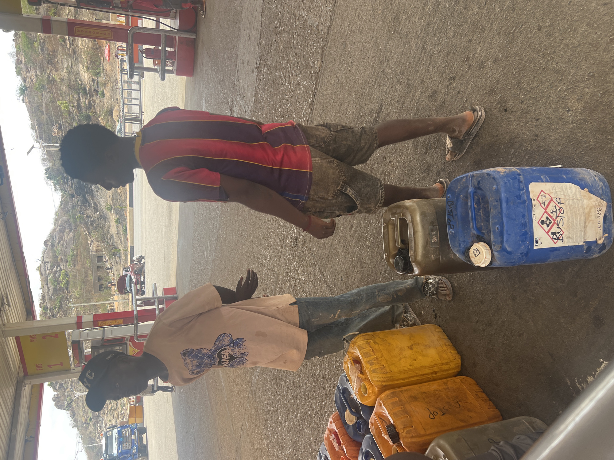 The image depicts an outdoor scene at what appears to be a fuel dispensing point or gas station in Jos, Nigeria, under bright daylight. Two young men are the primary subjects, alongside numerous plastic jerry cans.
The man on the left wears a light-colored t-shirt with a graphic (featuring a bear and possibly "NASA"), blue jeans, and a black baseball cap with "NYC" visible on the front. He is seen from his back and left profile, looking towards the left. The man on the right wears a red and black striped shirt and denim shorts, also viewed from his back and right profile, looking to the left. Both men wear sandals or slides.
In the foreground and around the men, a collection of plastic jerry cans in various colors, including yellow, orange, blue, and dark green, are stacked on the concrete ground. One blue jerry can features a label with hazard pictograms (flame, corrosive, exclamation mark) and text including "ACTICIDE HF", "THOR", "BUSTOP", and "UN 3265". Other containers have partial text visible, such as "BASTOPO", "BUSTOP", and "25 L".
In the background, red and white fuel pumps or dispensers are partially visible; one clearly displays "PMS 1" and "PMS 2", while another has "NOSHOKIA" on its side. A red fire extinguisher is also seen near a pump. Behind the station structure, a distinct rocky, hilly landscape with sparse vegetation stretches under a bright, clear sky.
The scene suggests an activity related to the acquisition or distribution of fuel or other liquids, likely in bulk, given the numerous containers. The men appear to be engaged in handling or waiting with their jerry cans. The bright lighting indicates midday or afternoon.