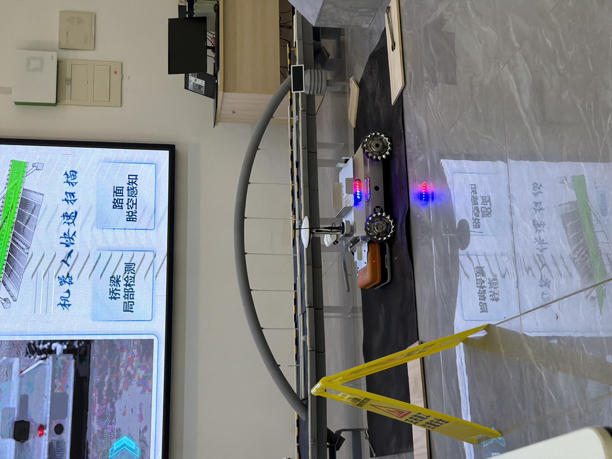 A mobile robot with flashing blue and red lights is positioned on a black mat in front of a miniature bridge exhibit. The robot has four wheels with intricate treads and appears to be actively engaged in some form of demonstration or testing. To the left of the robot, a yellow A-frame sign with a red triangle warning symbol is placed on the floor. The background features a large screen displaying information in Chinese characters, depicting a robot performing a quick scan of a bridge and road surface. A computer monitor and other electronic equipment are visible on a desk in the background. The floor is reflective, showing the lights from the robot and the signage. This scene is likely set within an educational or research facility in Jiangjin, China, given the Chinese text on the screen and the technological exhibit.
