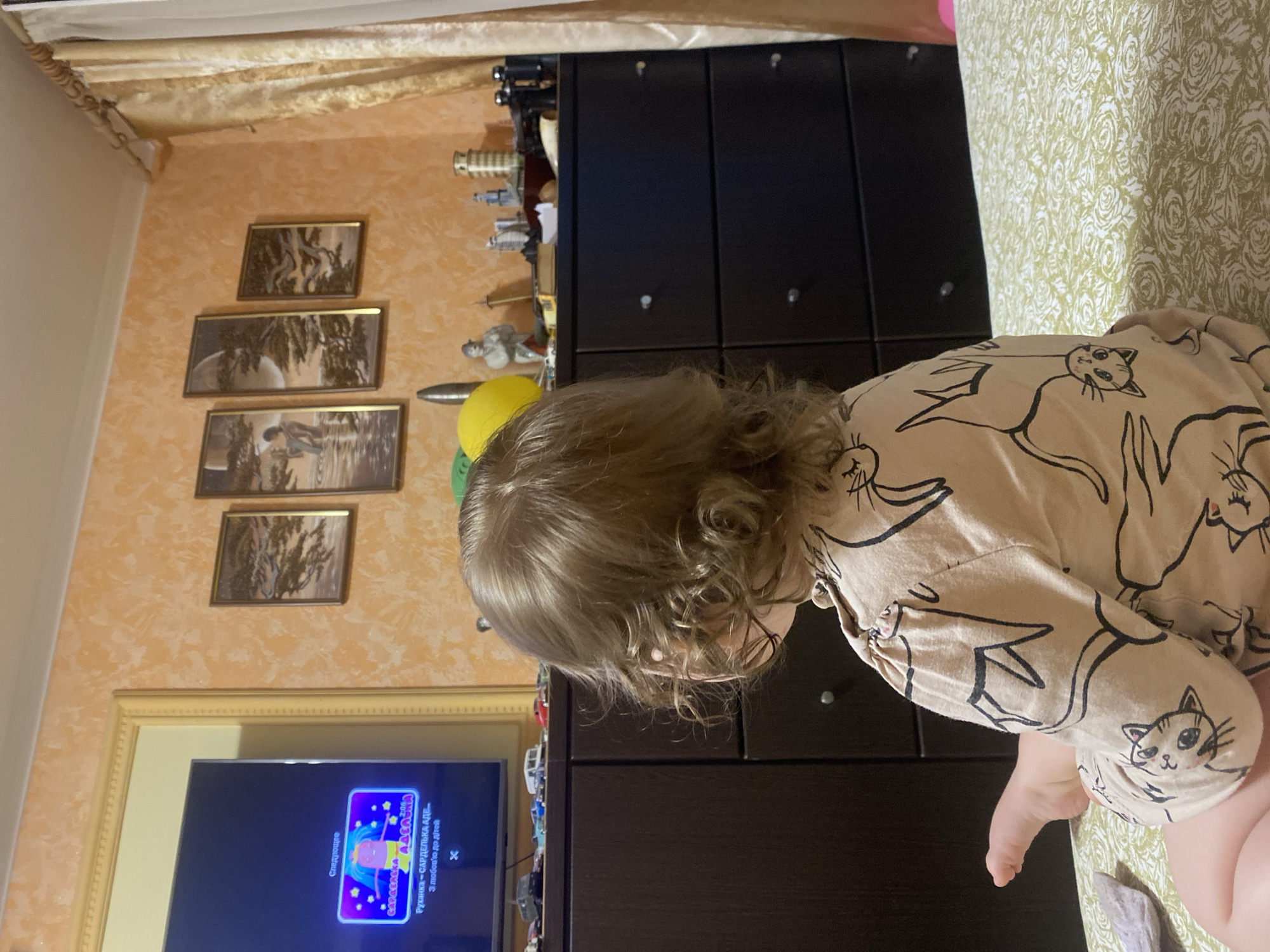 A young child with curly blonde hair sits facing away from the camera, engrossed in watching a television screen in a room with orange walls. The child is wearing a light-colored onesie adorned with a pattern of black cartoon cats. The television displays a bright purple graphic with text in Russian. To the left of the television, a dark brown dresser is visible, with a collection of toy cars arranged on top. Above the dresser, a series of framed artworks depicting natural scenes hang on the wall. To the right of the dresser, a yellow balloon and various decorative objects, including binoculars and miniature buildings, are placed. A sheer, gold-colored curtain hangs on the far right side of the image. The child is positioned on a bed covered with a light green, floral-patterned sheet.