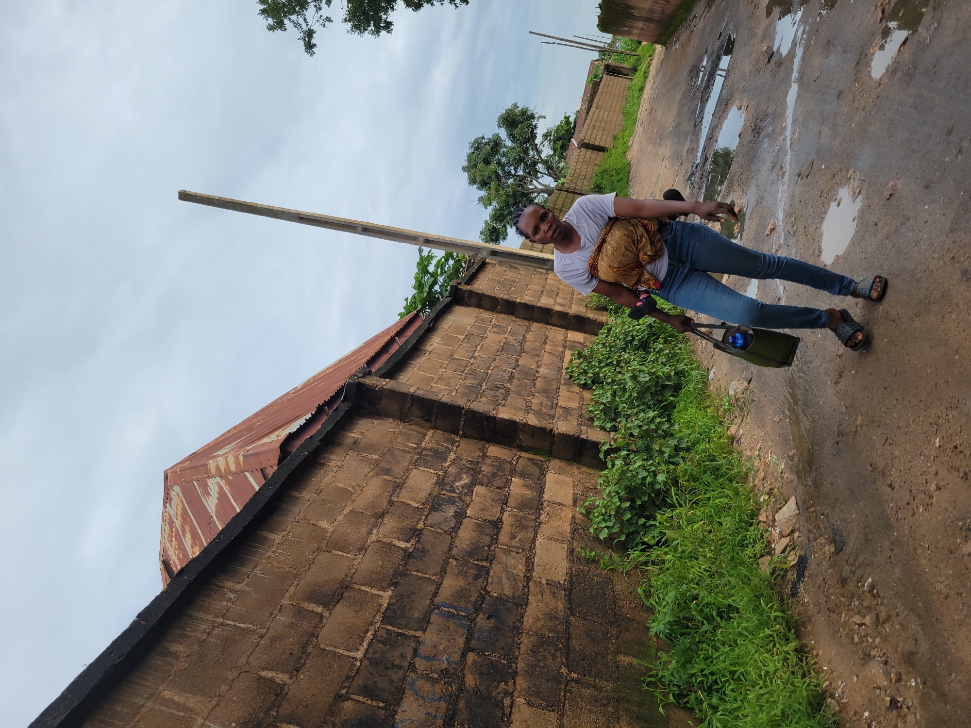 In Jos, Nigeria, a woman stands on a wet, unpaved road, her body angled towards the camera. She wears a light-colored t-shirt, blue jeans, and dark sandals. A patterned fabric wraps around her torso, securing a baby to her back, though the baby is not visible. In her left hand, she carries a dark green bag with a blue water bottle inside. The road is uneven and dotted with puddles, reflecting the overcast sky. To the left of the road is a tall, rough-hewn brick wall with a corrugated metal roof showing signs of rust. Lush green foliage grows along the base of the wall. A tall, weathered utility pole stands behind the woman, leaning slightly. In the background, more brick walls and trees line the road, suggesting a residential area. The overall atmosphere is subdued, with the cloudy sky contributing to a muted color palette.